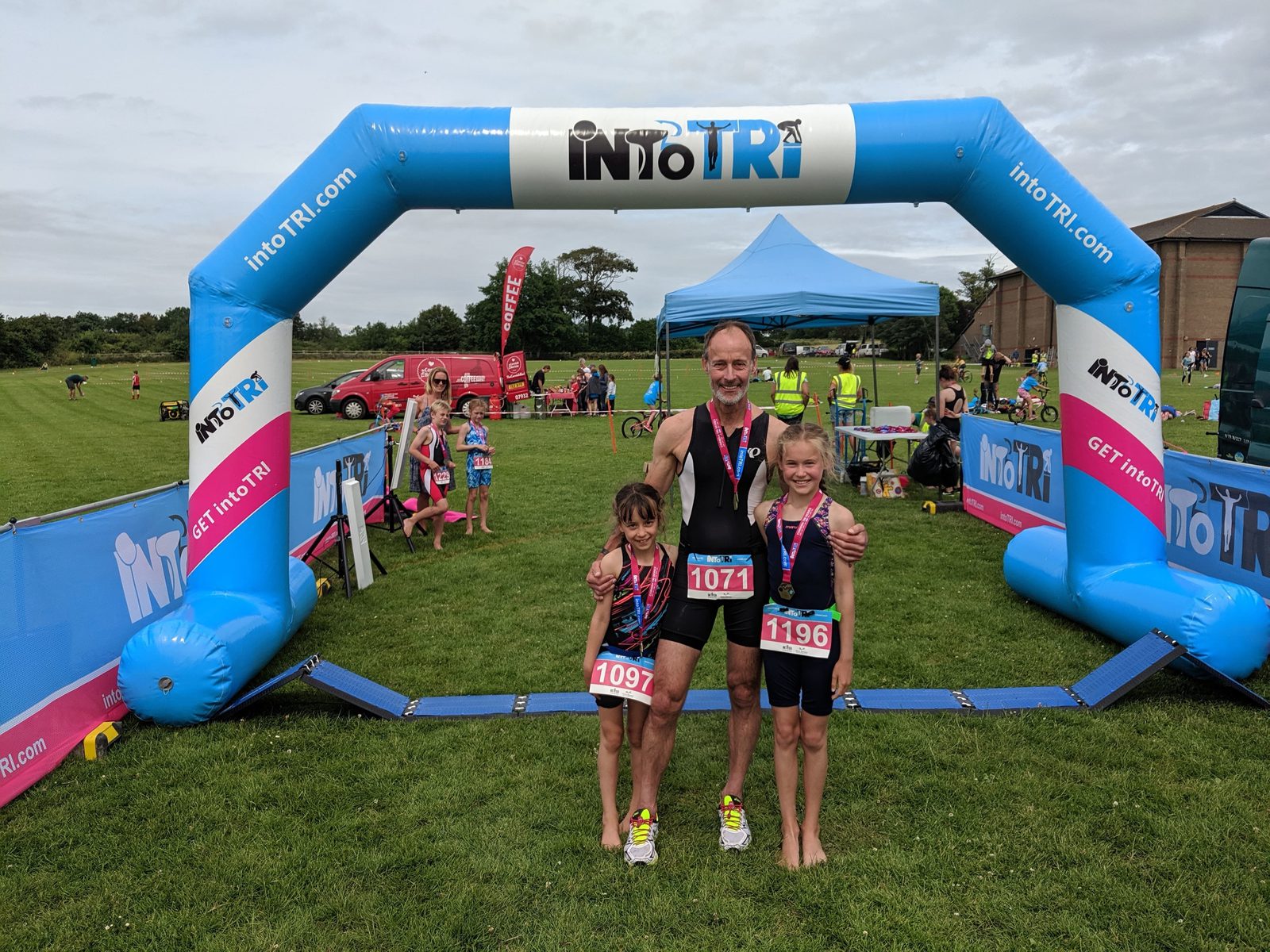 A man and two children, wearing athletic gear and medals, stand together and smile under a blue inflatable archway that reads "into TRI." They are standing on green grass with event tents, banners, and other participants in the background. The children hold small trophies.