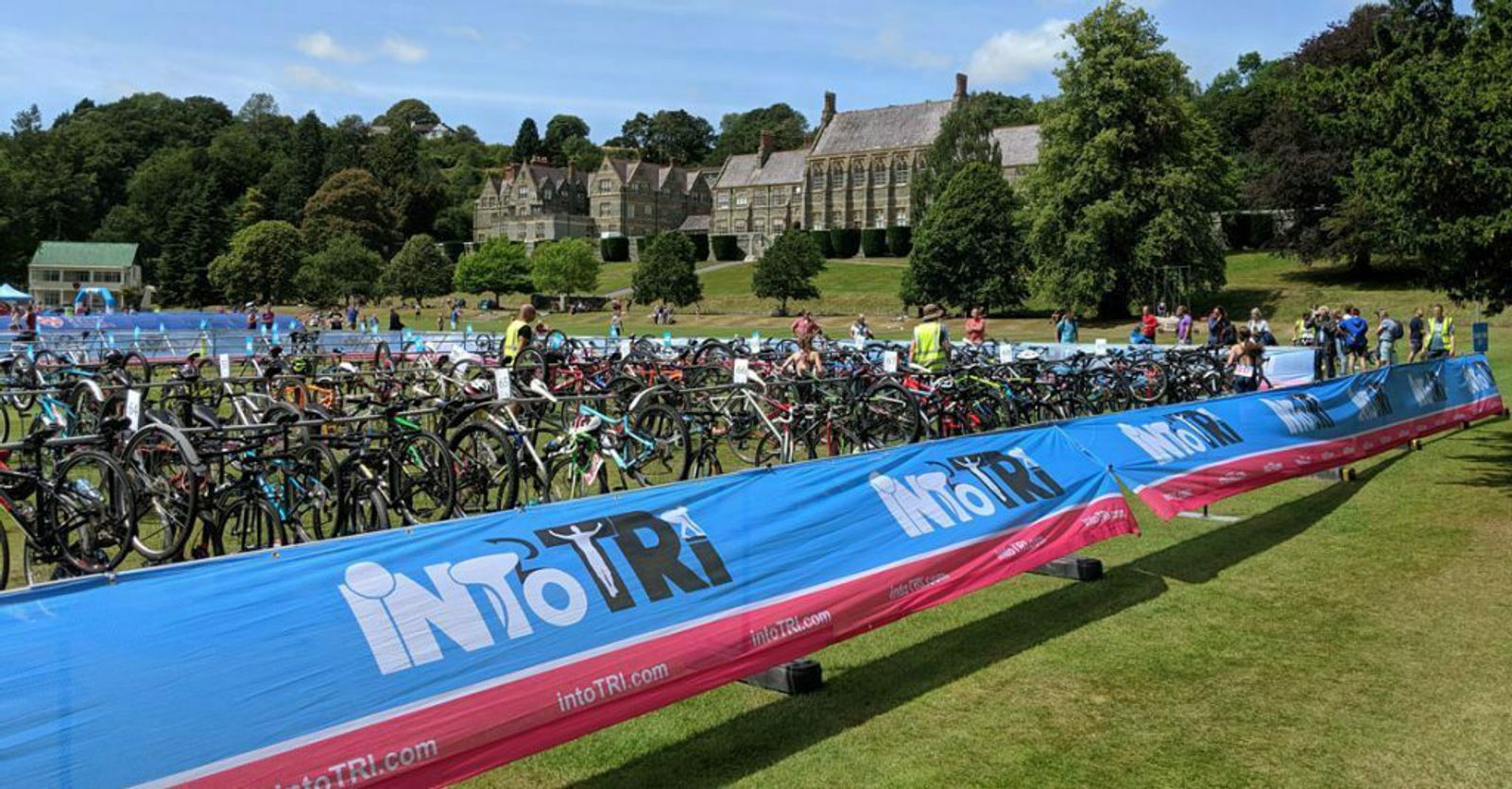 A large outdoor area with numerous bicycles parked in rows, surrounded by blue and red barriers labeled "intoTRI." Participants and staff are visible in the background, and a historic building is set amidst lush greenery in the distance.
