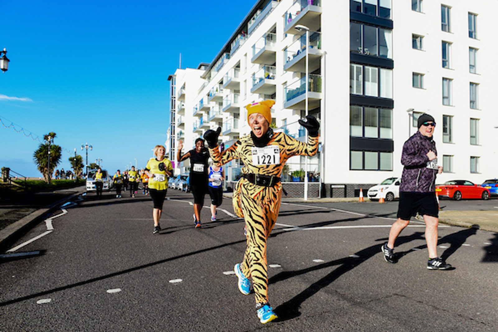 A runner dressed in a tiger costume with bib number 1221 waves cheerfully during a race on a sunny day. Several other runners are visible in the background. The race takes place on a paved road alongside modern white buildings and palm trees.