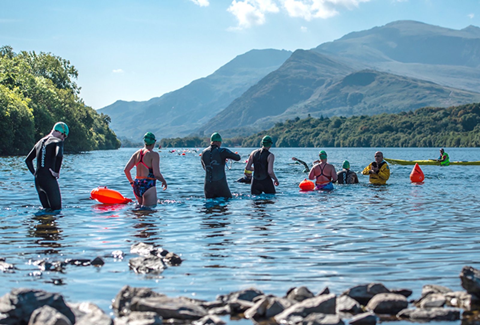 A group of people in swimsuits and green swim caps stand in a lake surrounded by mountains and greenery. Some are holding orange floatation devices, and a few kayakers are visible in the background under a sunny, blue sky.