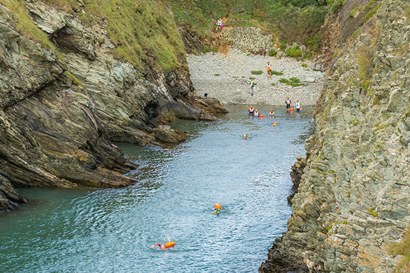 People swimming and wading in a narrow river between steep, rocky cliffs. Some individuals are on a small pebble beach area at the top of the river bank. The water is clear, and the scene is surrounded by lush greenery.
