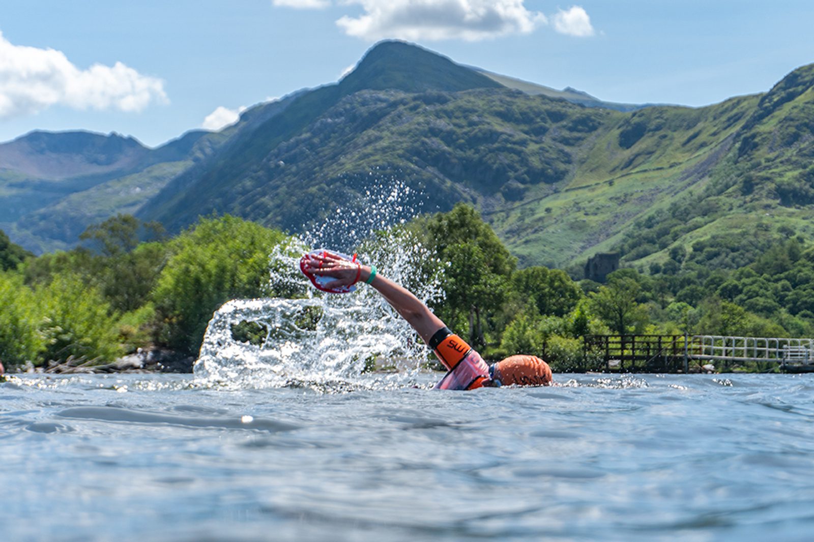 A swimmer in a bright orange cap and matching wristbands swims in a clear lake surrounded by lush green hills and mountains under a blue sky with scattered clouds. The swimmer's arm is extended mid-stroke, and water splashes around.