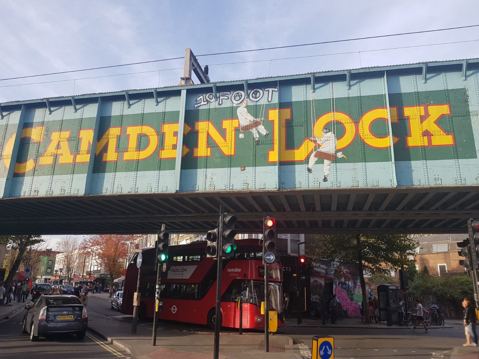 A bustling street scene in Camden, London, featuring the iconic Camden Lock railway bridge with its distinctive blue and yellow signage. The area is busy with pedestrians, cars, and a red double-decker bus. Traffic lights and buildings are visible in the background.