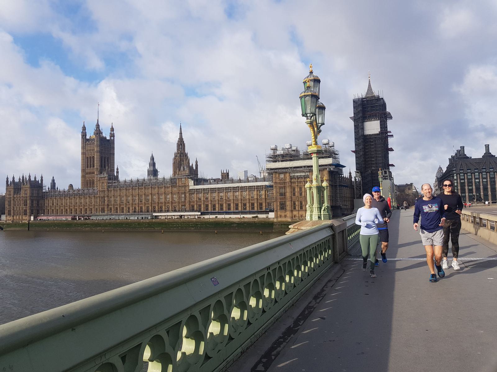 A group of people are jogging across Westminster Bridge in London. The Palace of Westminster and the Elizabeth Tower, partially covered in scaffolding, are visible in the background. The sky is partly cloudy, and the River Thames flows alongside the bridge.