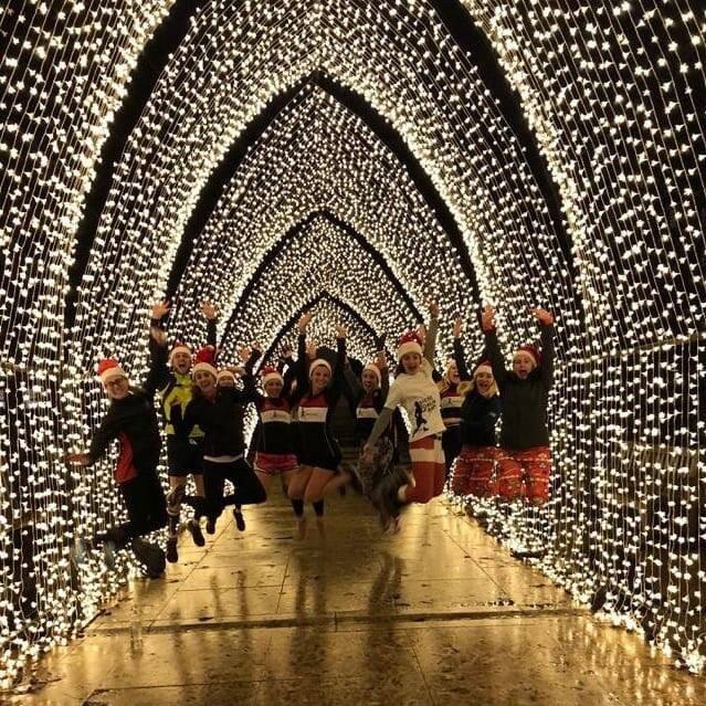 A group of people in festive clothing and Santa hats jump in unison under an archway adorned with thousands of twinkling Christmas lights, creating a tunnel of illuminated arches. The ground is wet, reflecting the dazzling lights above.
