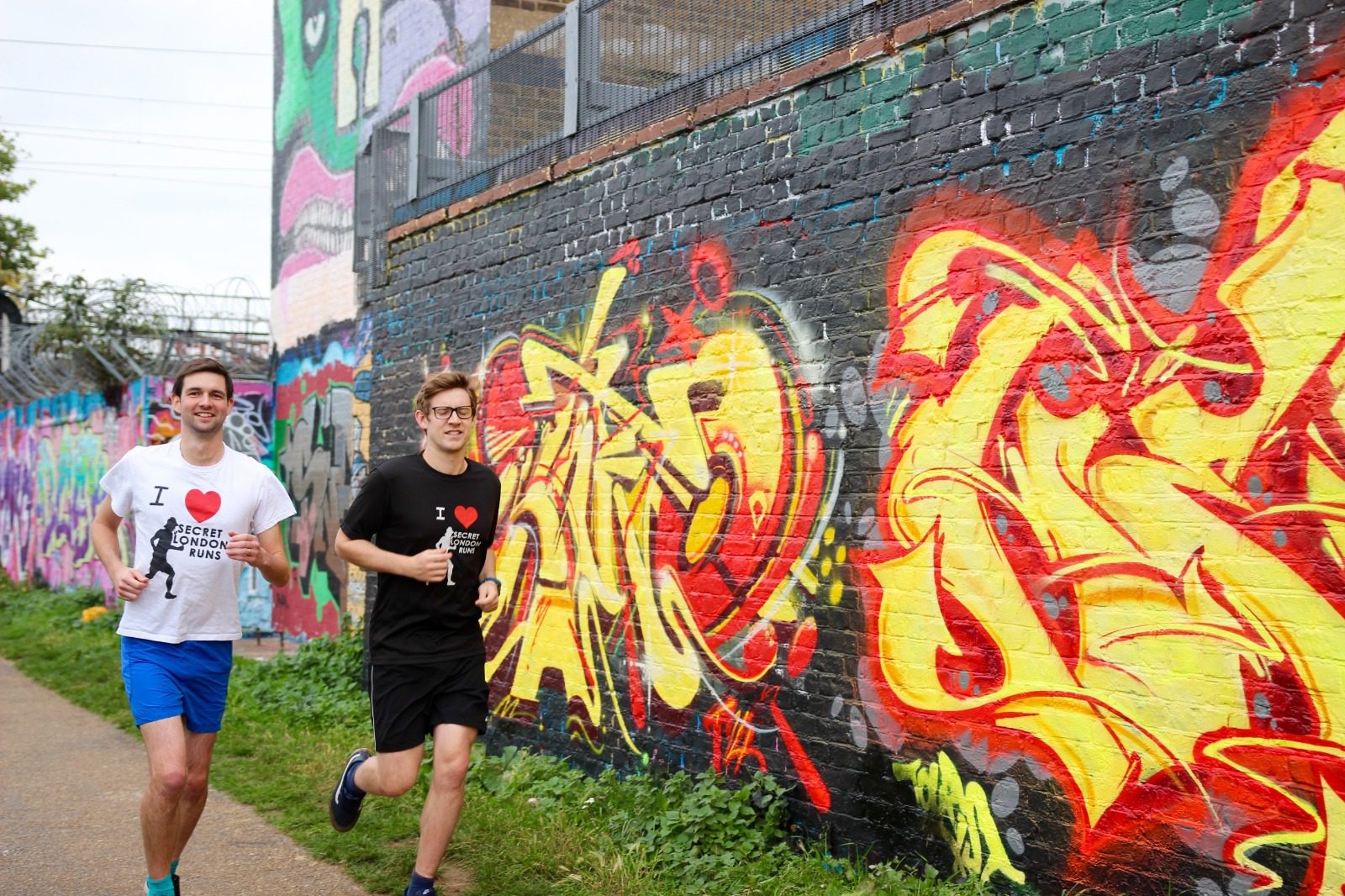 Two young men are jogging past a colorful graffiti-covered wall. Both are smiling, wearing T-shirts with "I ❤️ Hackney", and one is in black while the other is in white. The vibrant graffiti features abstract designs and bright colors.