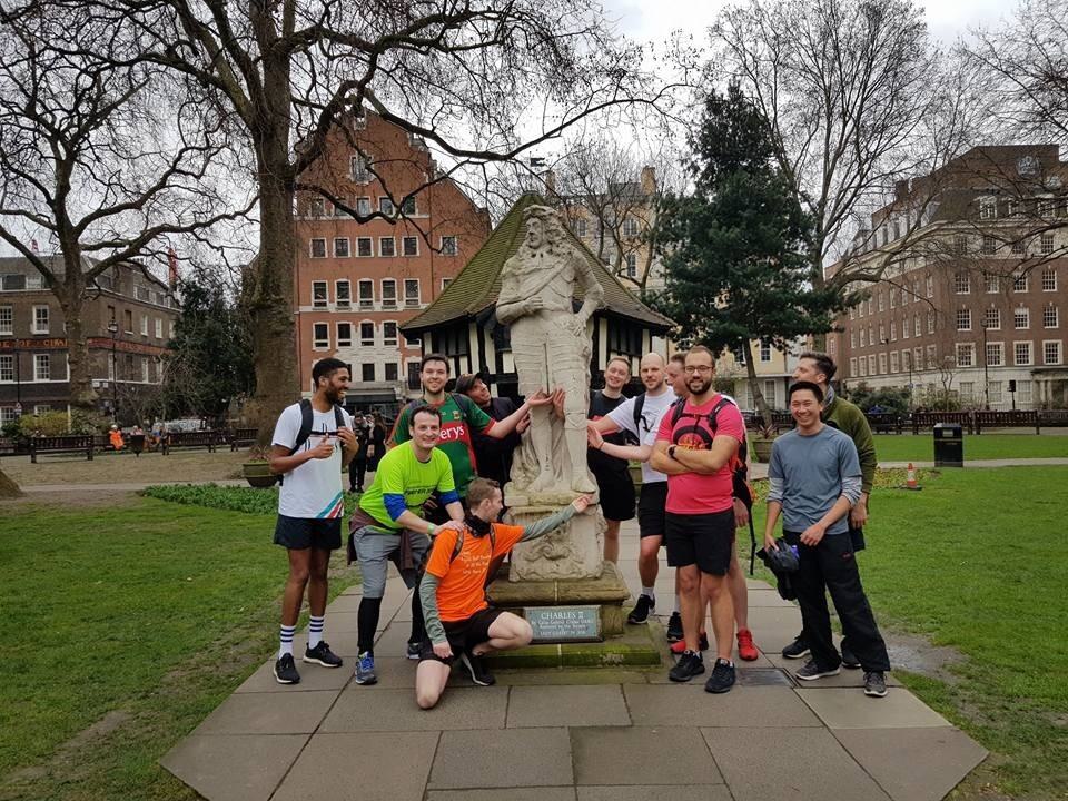 A group of twelve people, mostly men, gather around a stone statue of a figure in a park. Some people are wearing T-shirts and shorts, and others are wearing casual wear. The park has bare trees and buildings in the background. Everyone appears to be smiling or laughing.