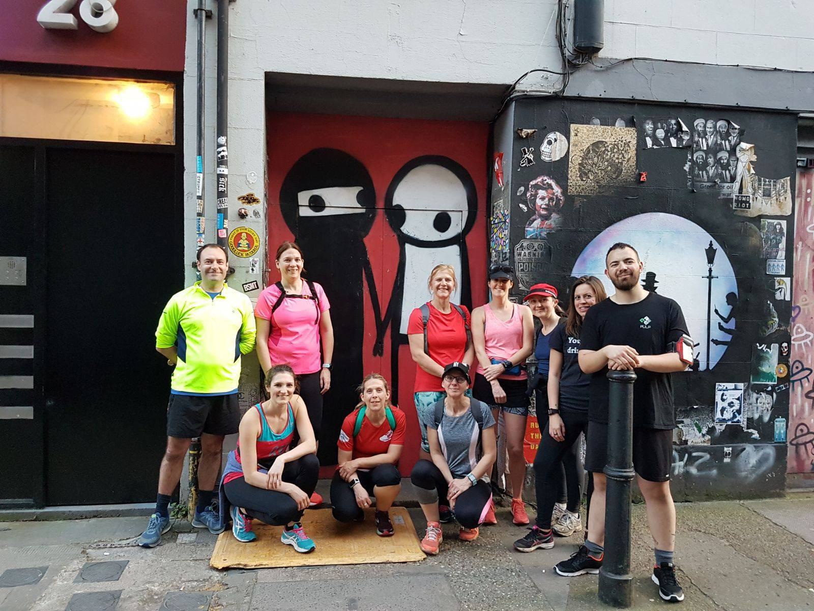 A group of twelve people, dressed in athletic gear, are posing in front of a mural featuring two stick figures. The background includes various pieces of street art on a gray wall. They appear to be runners, gathered near a building entrance marked with the number 28.