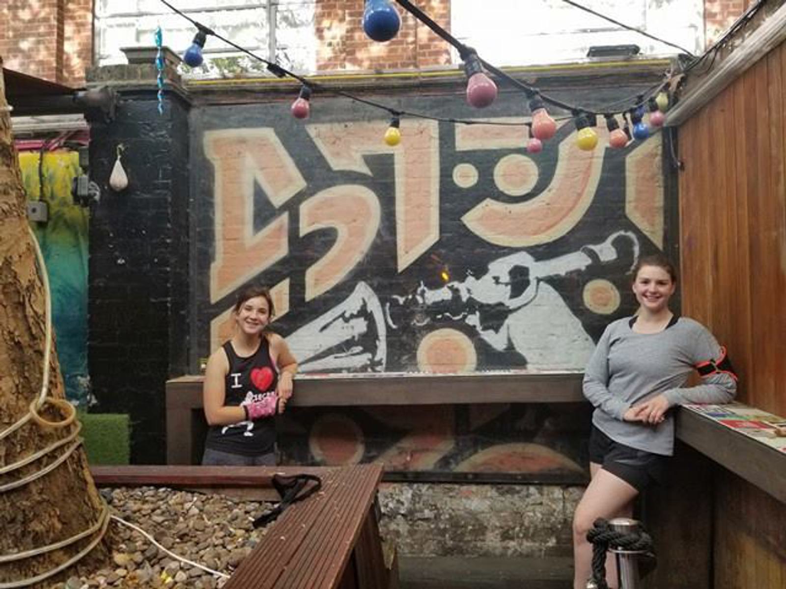 Two people are standing in front of a brick wall mural with abstract patterns and colors. String lights with colorful bulbs hang above them. The person on the left is wearing a tank top and smiling, while the person on the right leans on a counter with a relaxed expression.