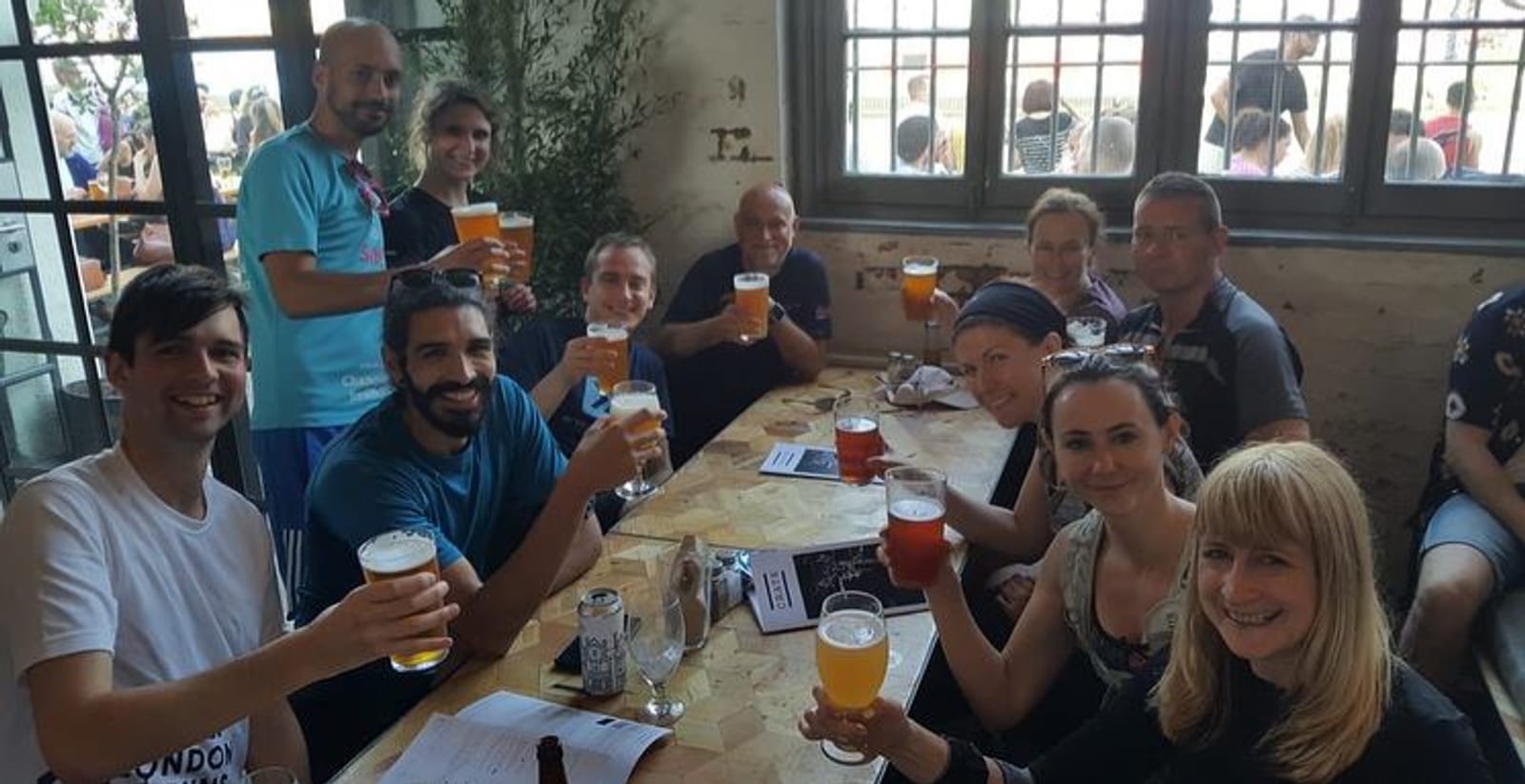 A group of people gathered around a long wooden table inside a rustic venue. They are raising their glasses and smiling at the camera, each holding a drink. The atmosphere appears cheerful and lively.