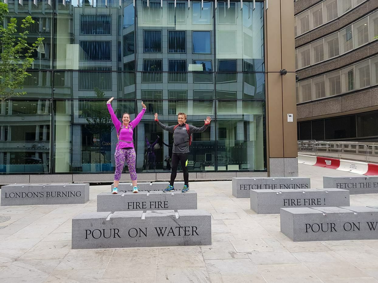 Two people are standing on stone platforms in front of a modern building. Each platform has engraved phrases like "FIRE FIRE" and "POUR ON WATER." Both individuals have their arms raised in the air, appearing joyful and energetic.
