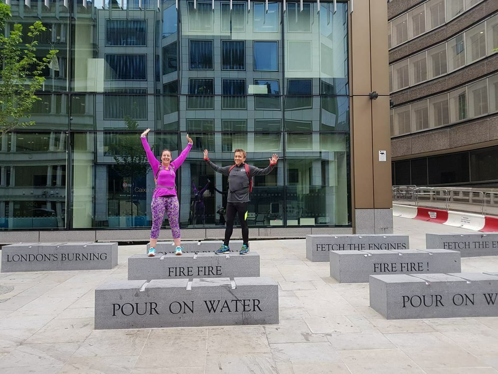 Two people are standing on stone platforms in front of a modern building. Each platform has engraved phrases like "FIRE FIRE" and "POUR ON WATER." Both individuals have their arms raised in the air, appearing joyful and energetic.