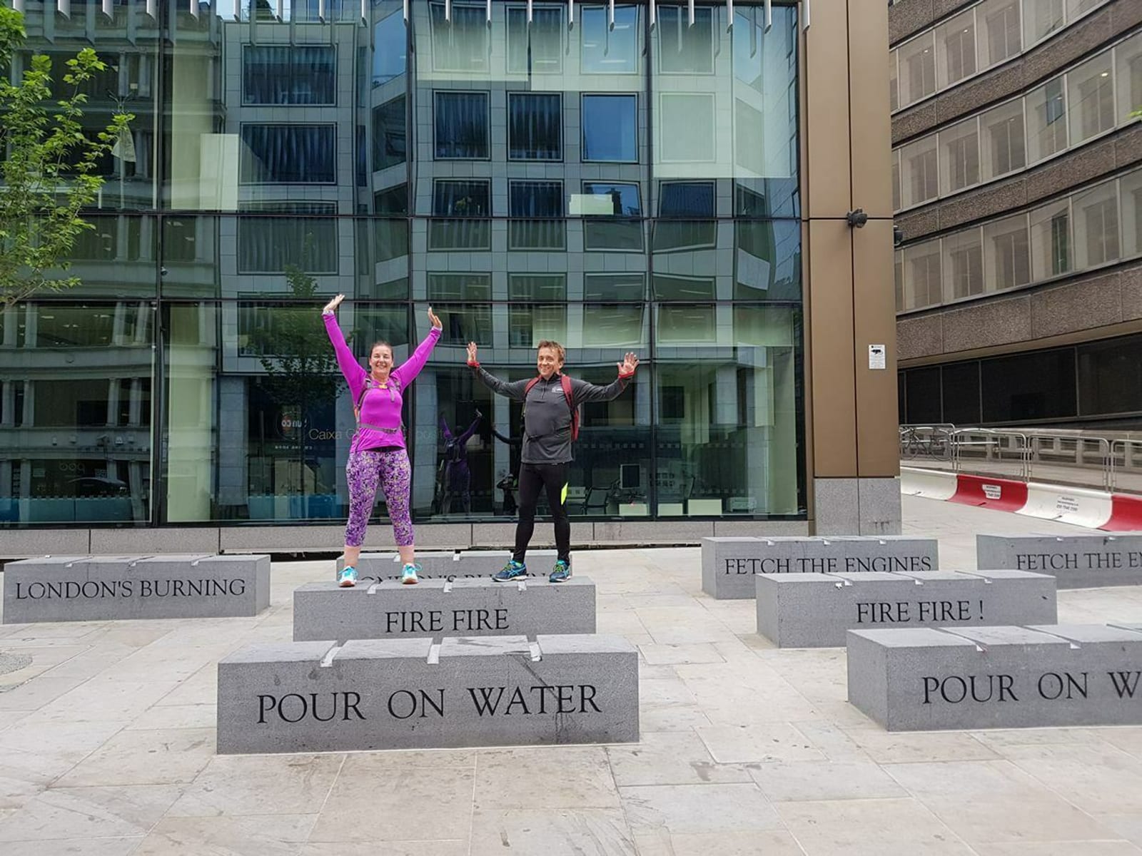 Two people are standing on stone blocks with engraved text, smiling and raising their arms in celebration. The blocks are located in an urban area with modern buildings in the background. The text on the blocks reads "LONDON'S BURNING," "FIRE FIRE," "POUR ON WATER," and "FETCH THE ENGINES.