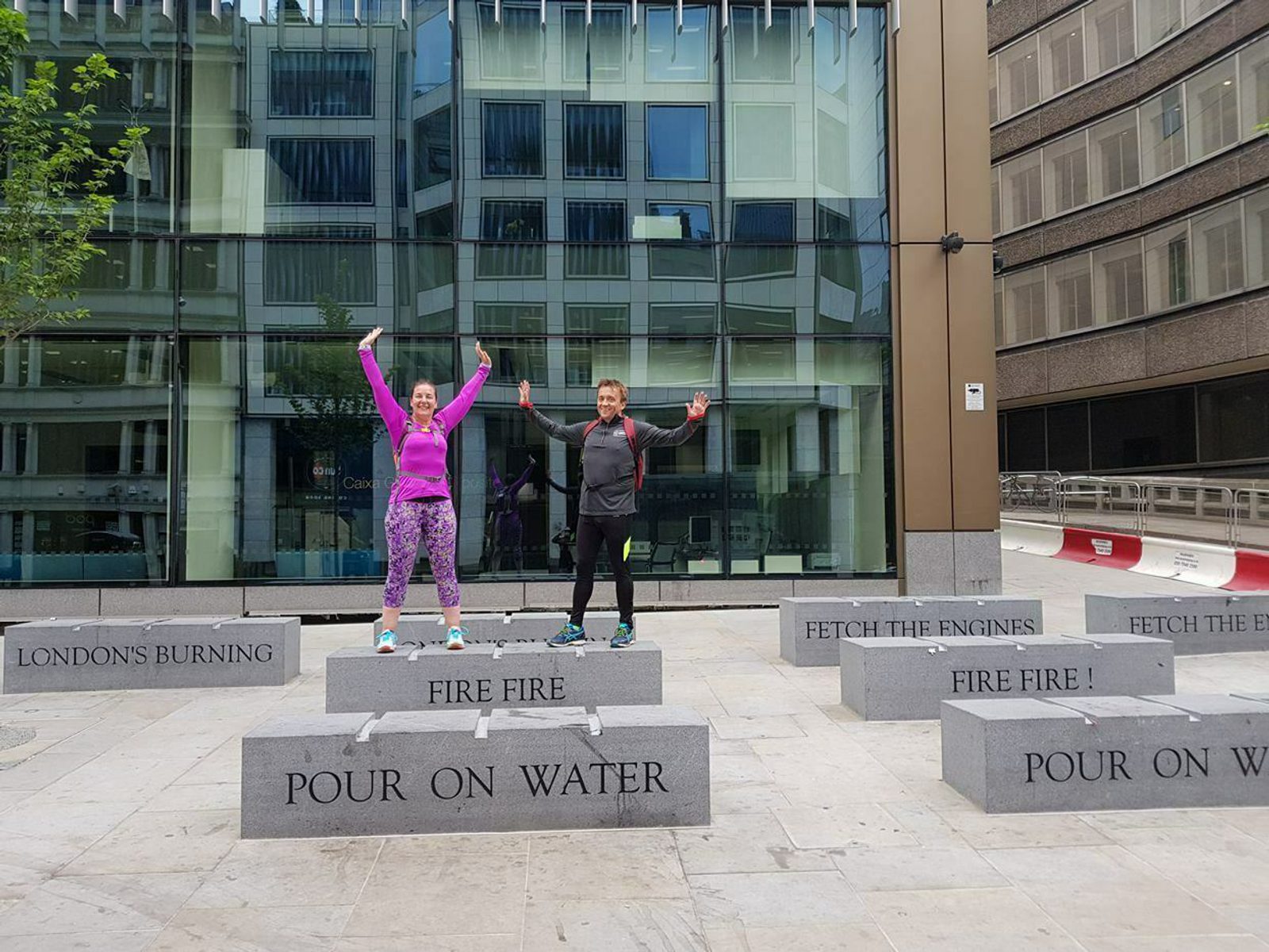 Two people are posing on a series of stone blocks with engraved text. One person, wearing a pink jacket, has their arms raised in the air. The other person, in a darker outfit, has their arms extended to the sides. A modern building serves as the backdrop.