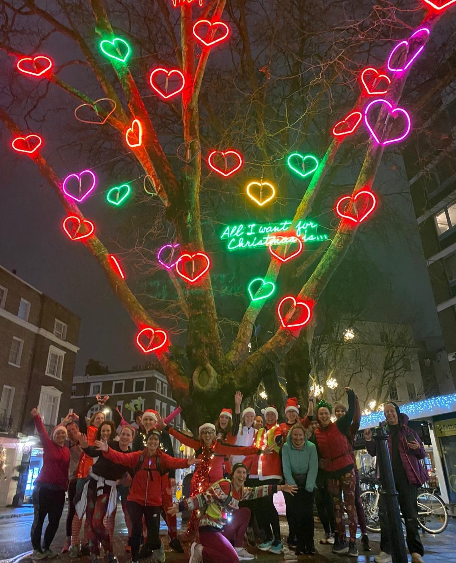 A group of people dressed in Christmas-themed outfits stand together on a city street at night. Behind them is a large tree adorned with colorful heart-shaped lights and a neon sign that reads, "All I want for Christmas is....." Buildings with holiday decorations surround them.