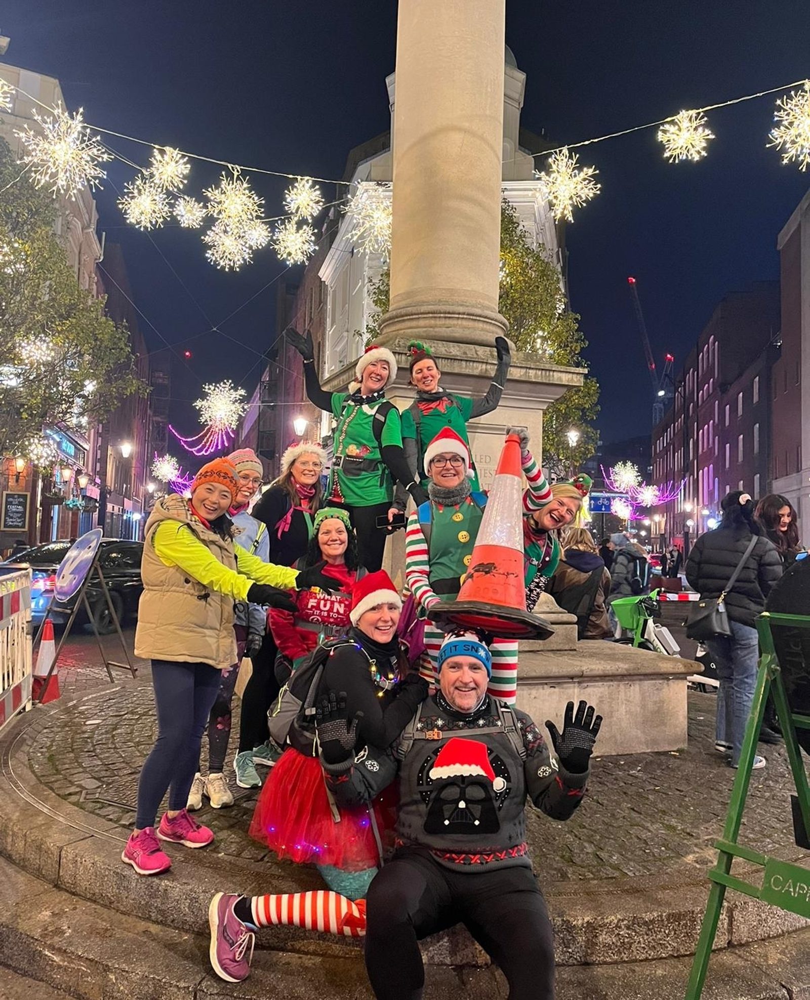 A group of people in festive attire poses happily around a large pillar at night. The street is decorated with lights and holiday ornaments. Some wear Santa hats, colorful costumes, and one holds a traffic cone. The atmosphere is lively and celebratory.