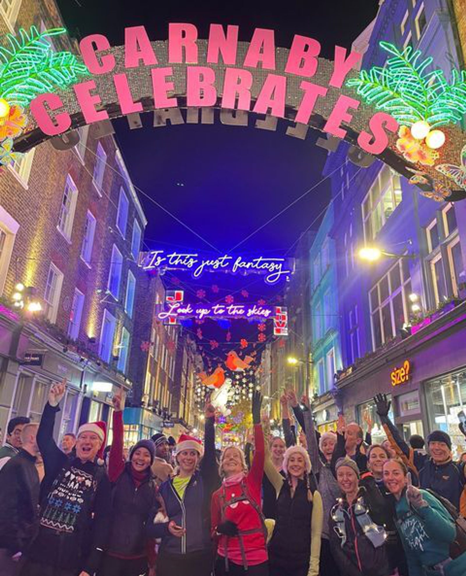 A group of people, some wearing Santa hats, are gathered on a festive street under a bright sign that reads "CARNABY CELEBRATES." The street is adorned with colorful lights and decorations, including a neon sign with the words "Is this just fantasy.