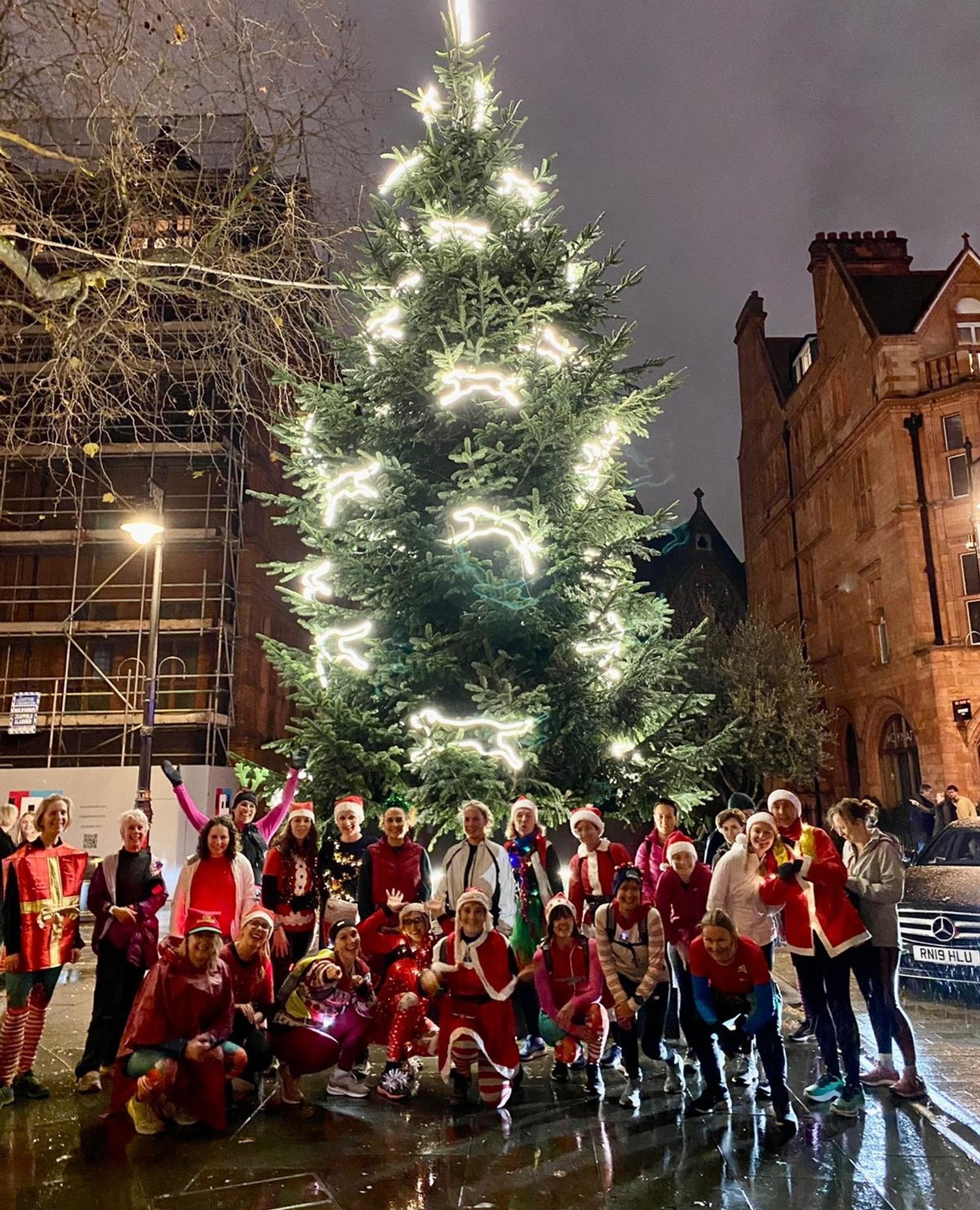 A group of people dressed in festive attire cheerfully pose in front of a large, illuminated Christmas tree at night. The tree is adorned with bright white lights, and the street is wet, reflecting the festive lights and creating a spirited holiday atmosphere.