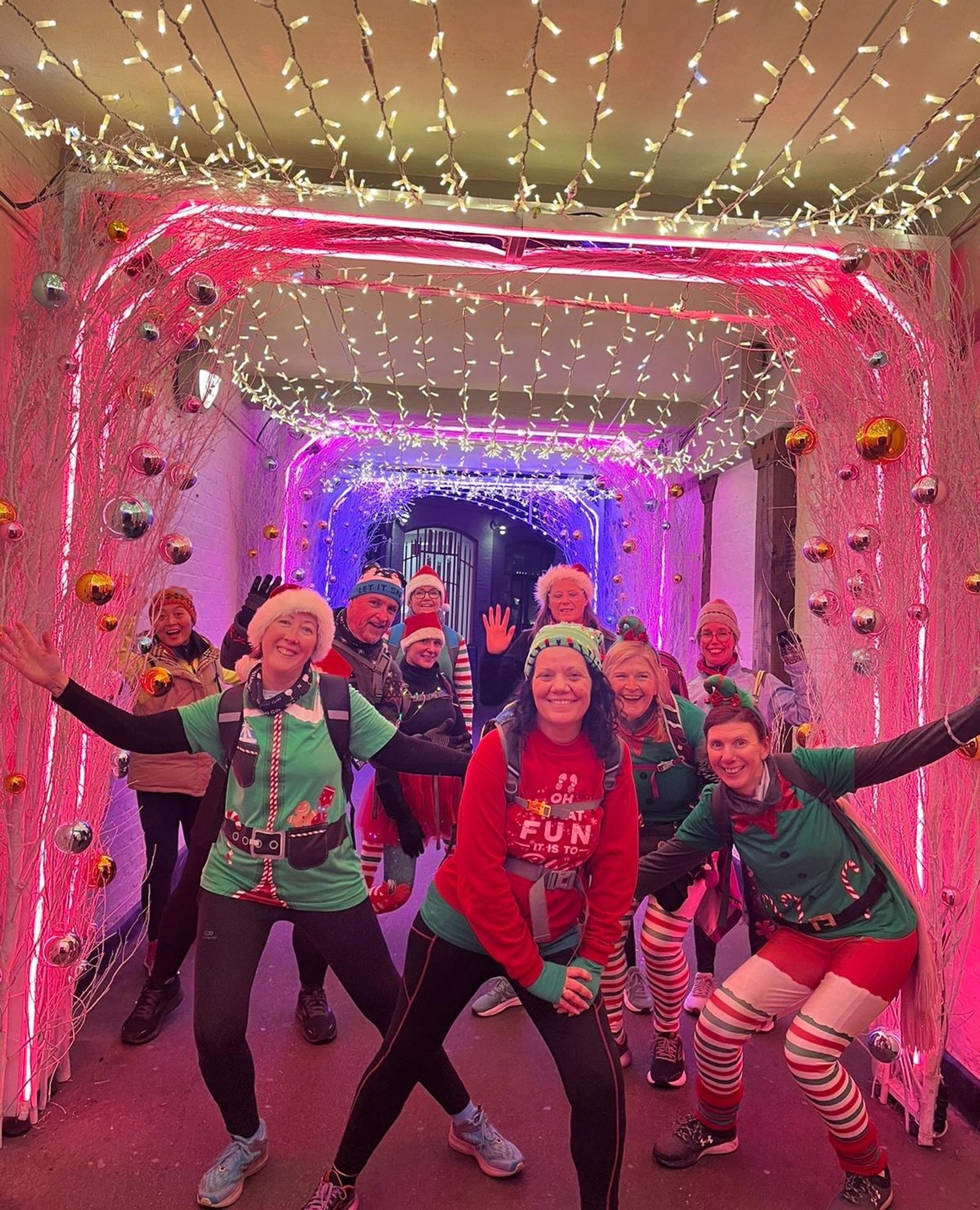 A group of people dressed in festive holiday outfits, including elf and Santa costumes, pose cheerfully under a tunnel adorned with colorful fairy lights and hanging ornaments. They appear to be in high spirits, enjoying the holiday season.