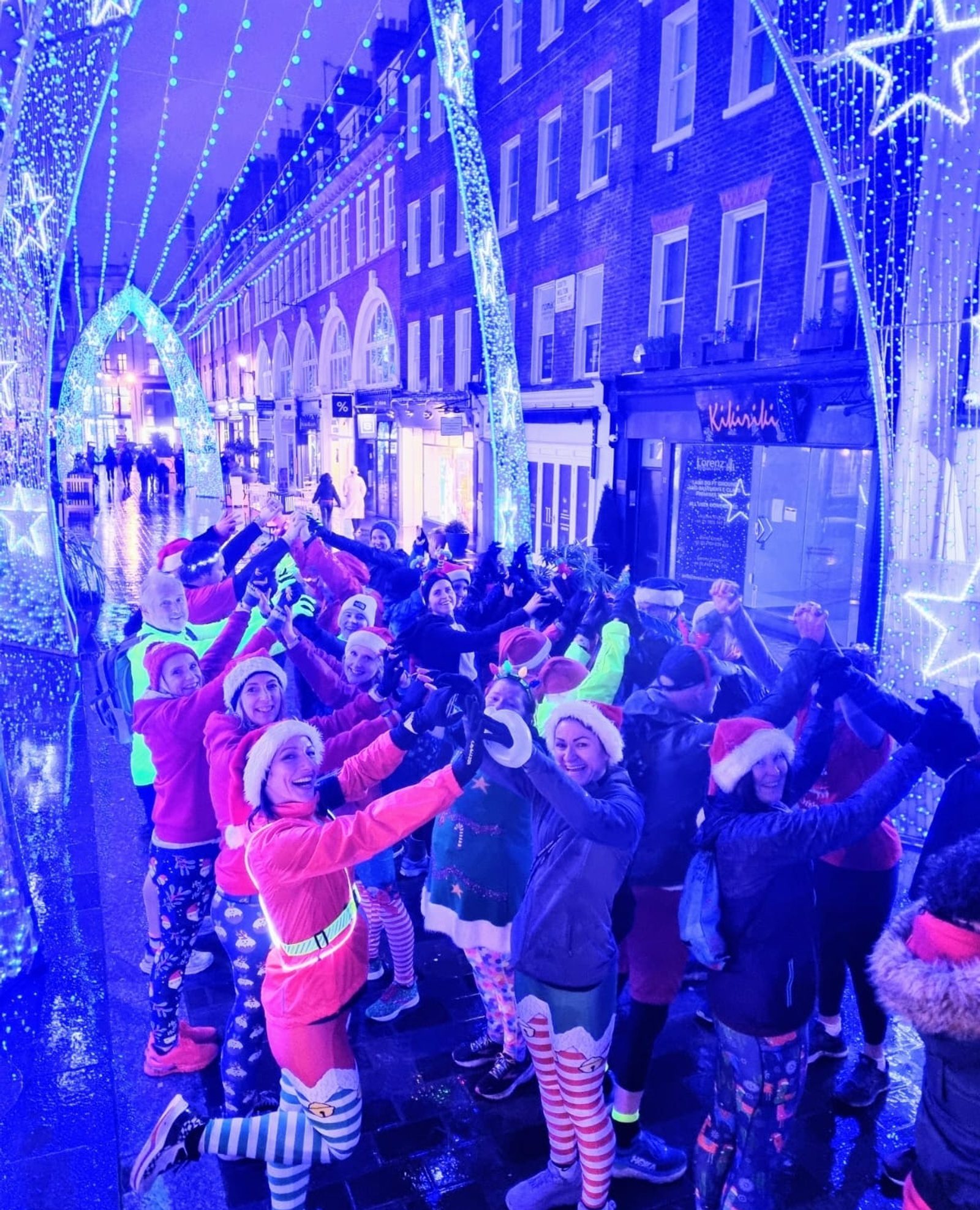 A group of people dressed in festive holiday clothing, including Santa hats and colorful outfits, are standing together under blue illuminated arches adorned with stars and lights. They are posing cheerfully on a street lined with shops at night.