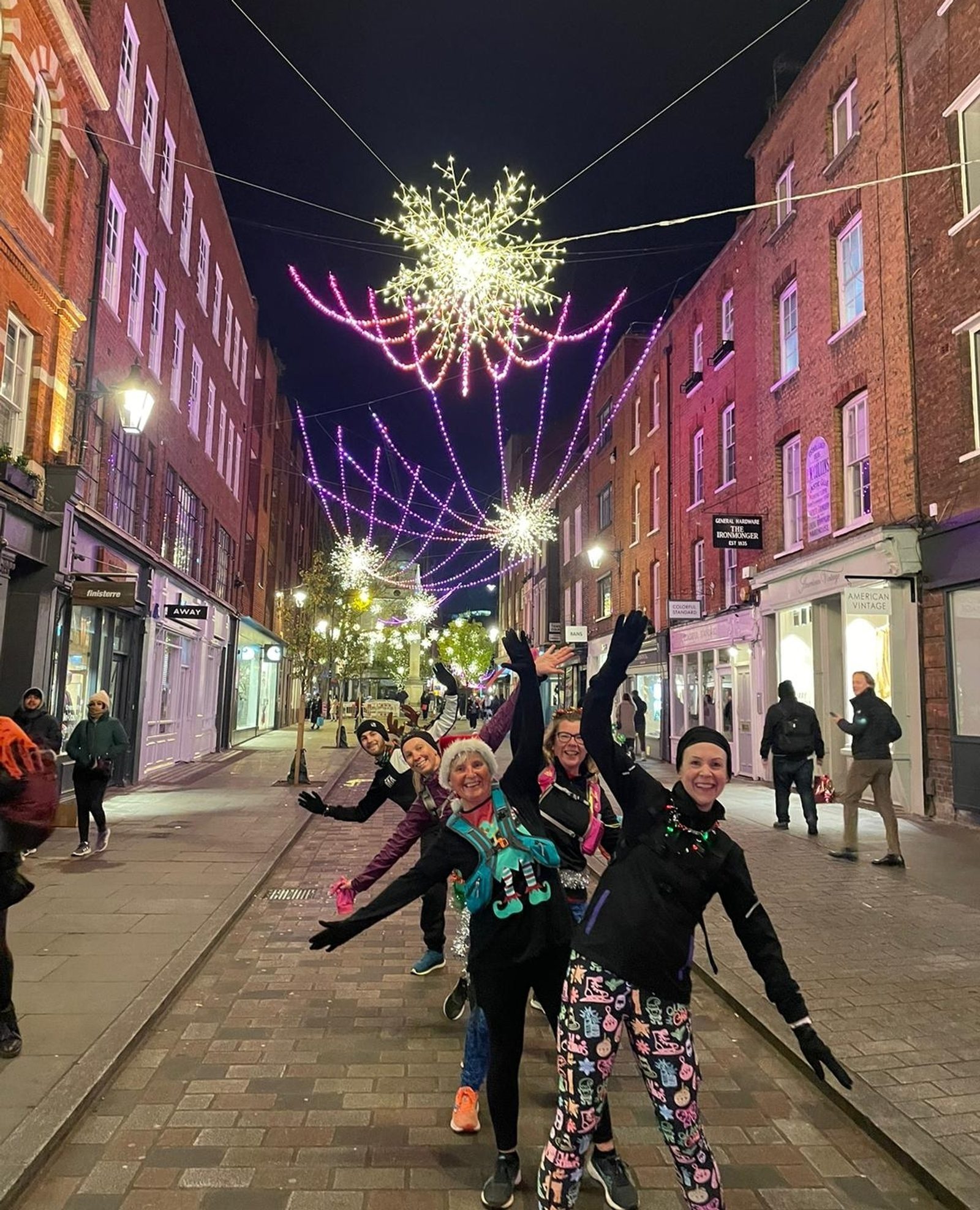 A group of people in festive attire pose energetically under illuminated decorations on a lively, brightly-lit street at night. The street is adorned with hanging light displays resembling snowflakes and ribbons, and the buildings on either side are aglow.
