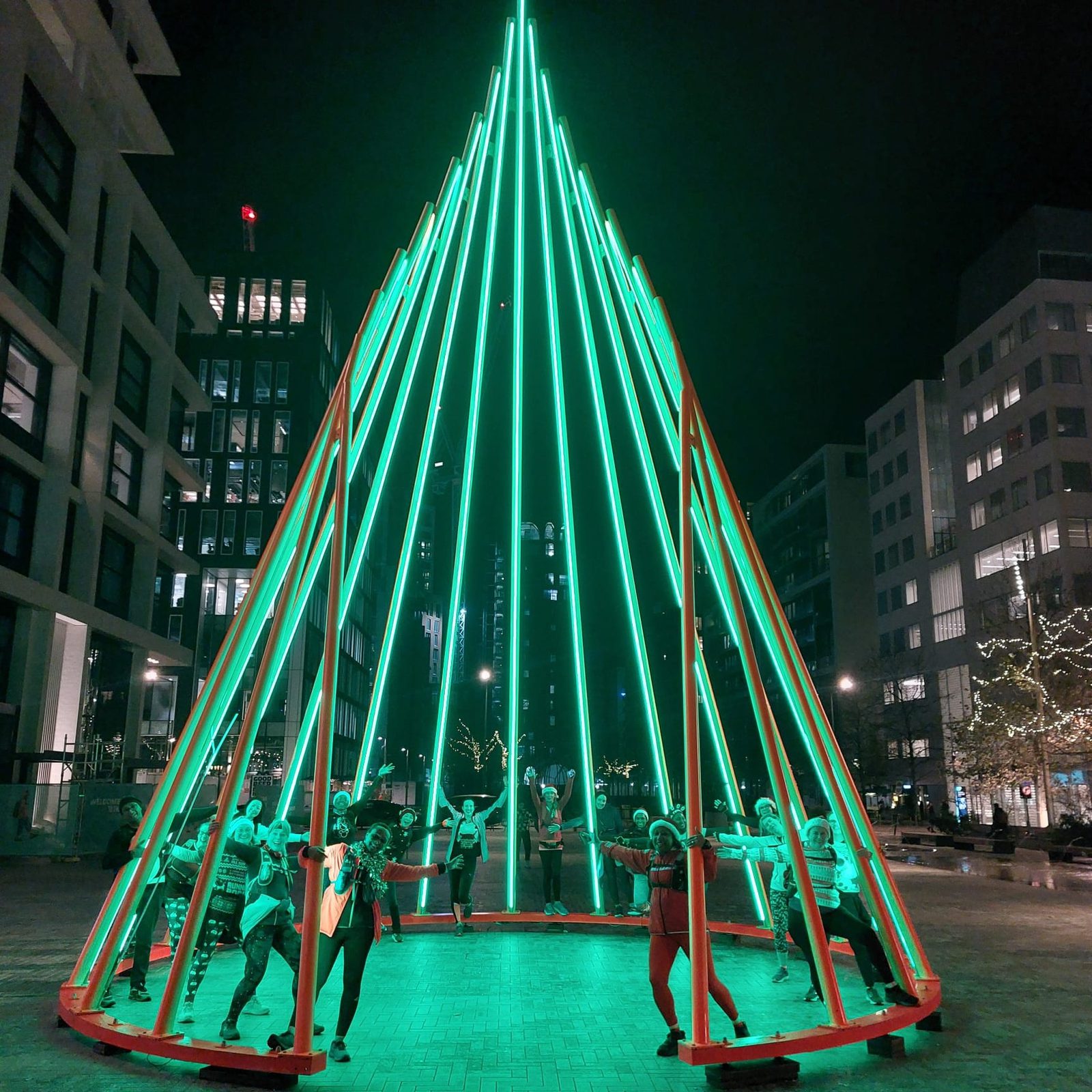 A group of people dressed in festive clothing gather around a large, triangular structure made of vertical green lights, resembling a Christmas tree, set against a backdrop of tall buildings with illuminated windows at night.
