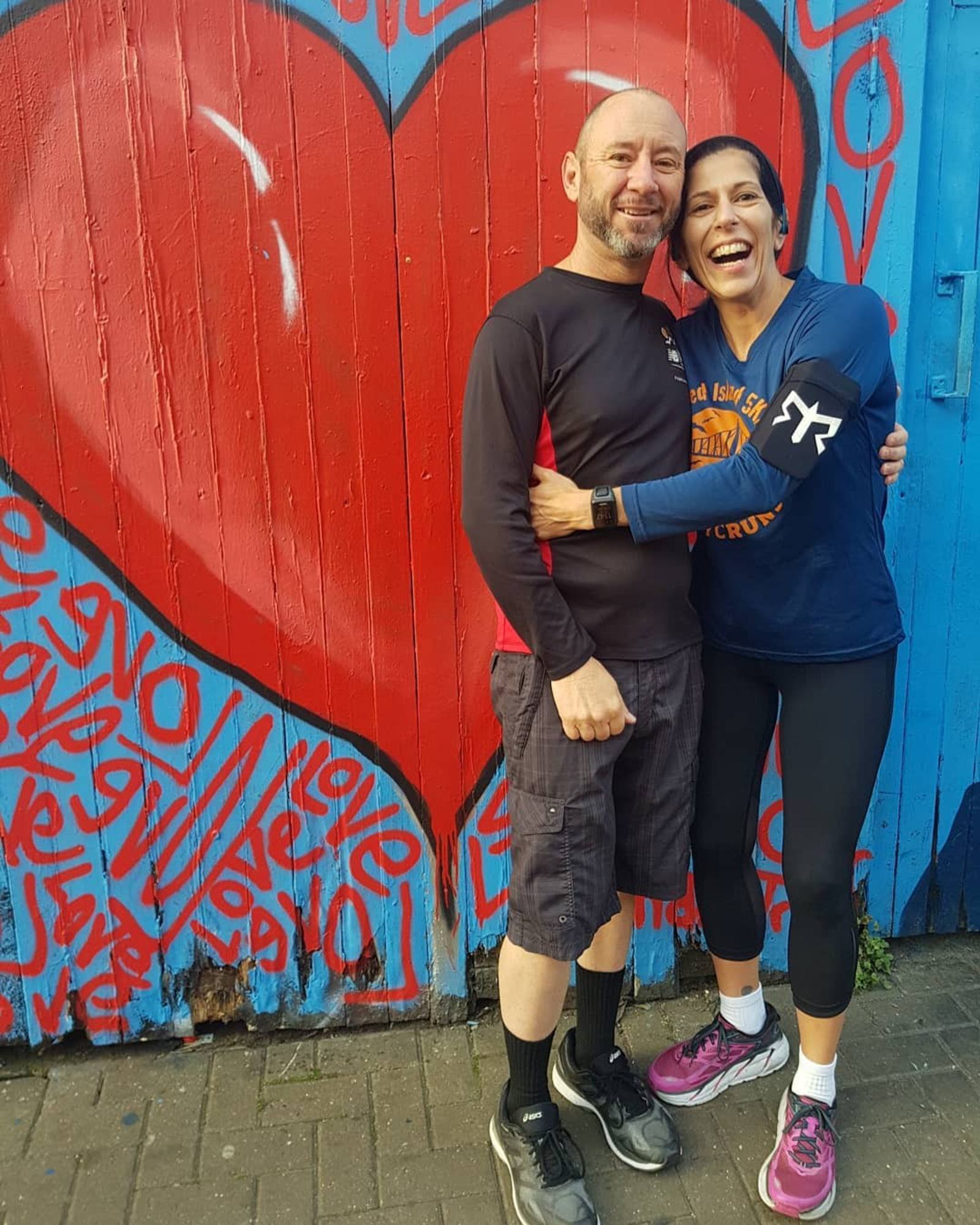 A man and a woman stand in front of a vibrant mural featuring a large red heart with blue and red background details. Both are dressed in athletic clothing and appear cheerful, with the woman laughing and the man smiling with his arm around her.