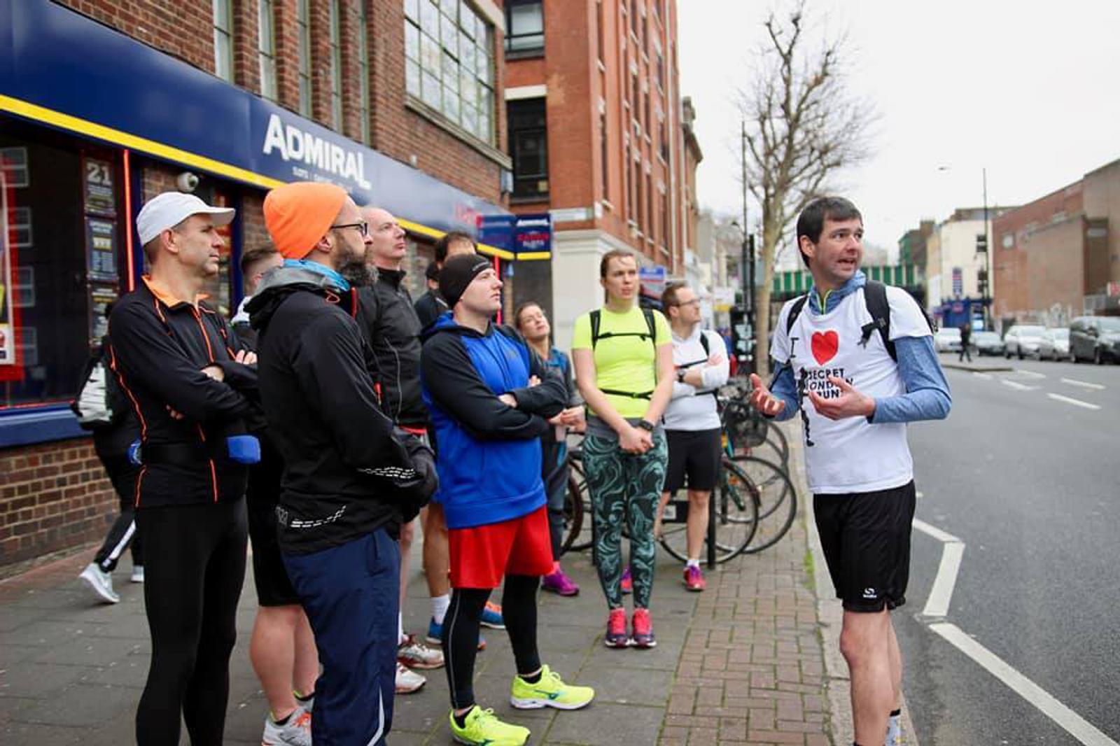 A group of runners is gathered on a city street, listening attentively to a person in a white T-shirt who appears to be giving instructions. They are dressed in various running gear, including jackets, shorts, and brightly colored athletic shoes.
