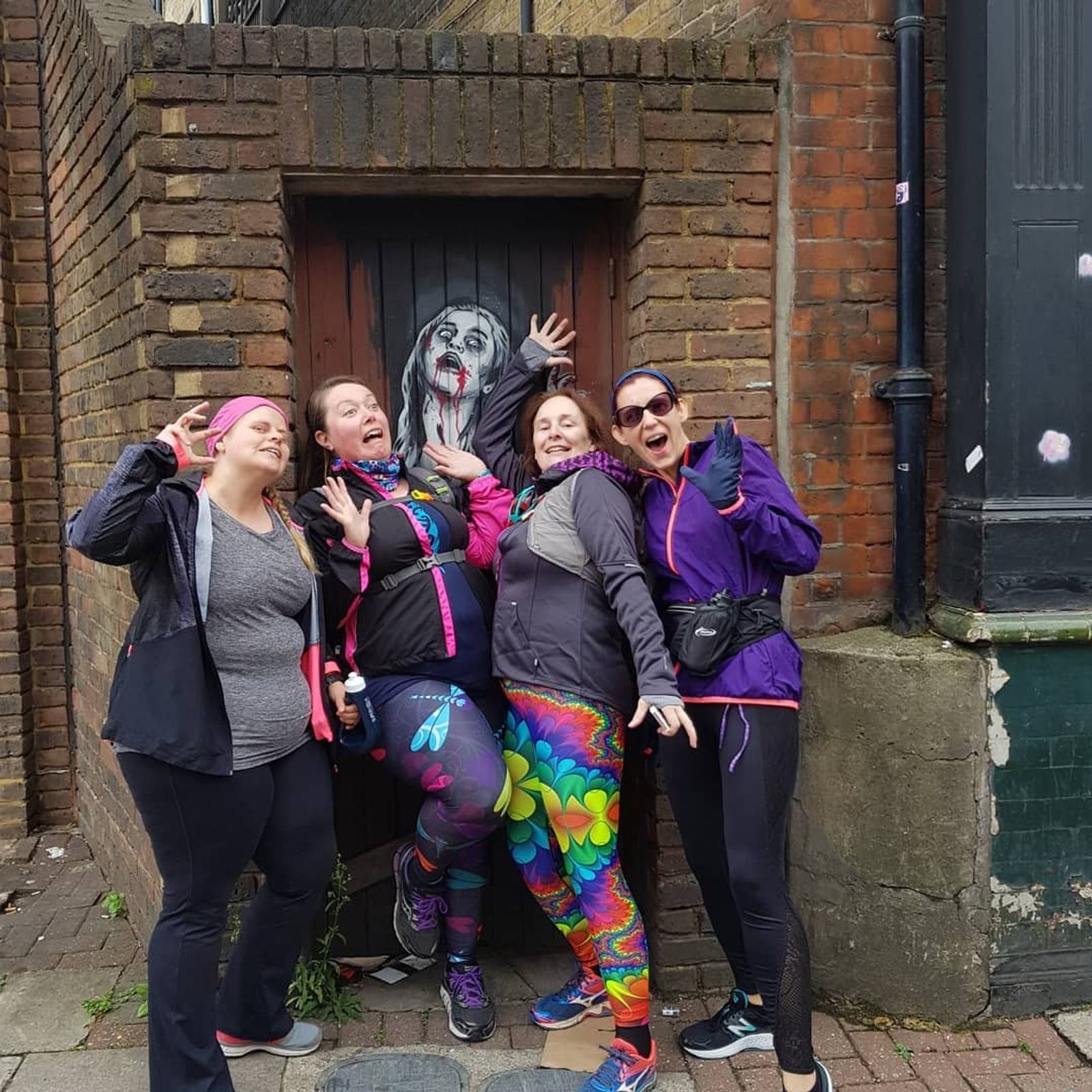 Four women strike playful poses in front of a mural depicting a spooky figure with a painted, distressed face and hands pressed against the door. They are dressed in colorful, casual athletic wear and appear to be having fun in the urban setting.