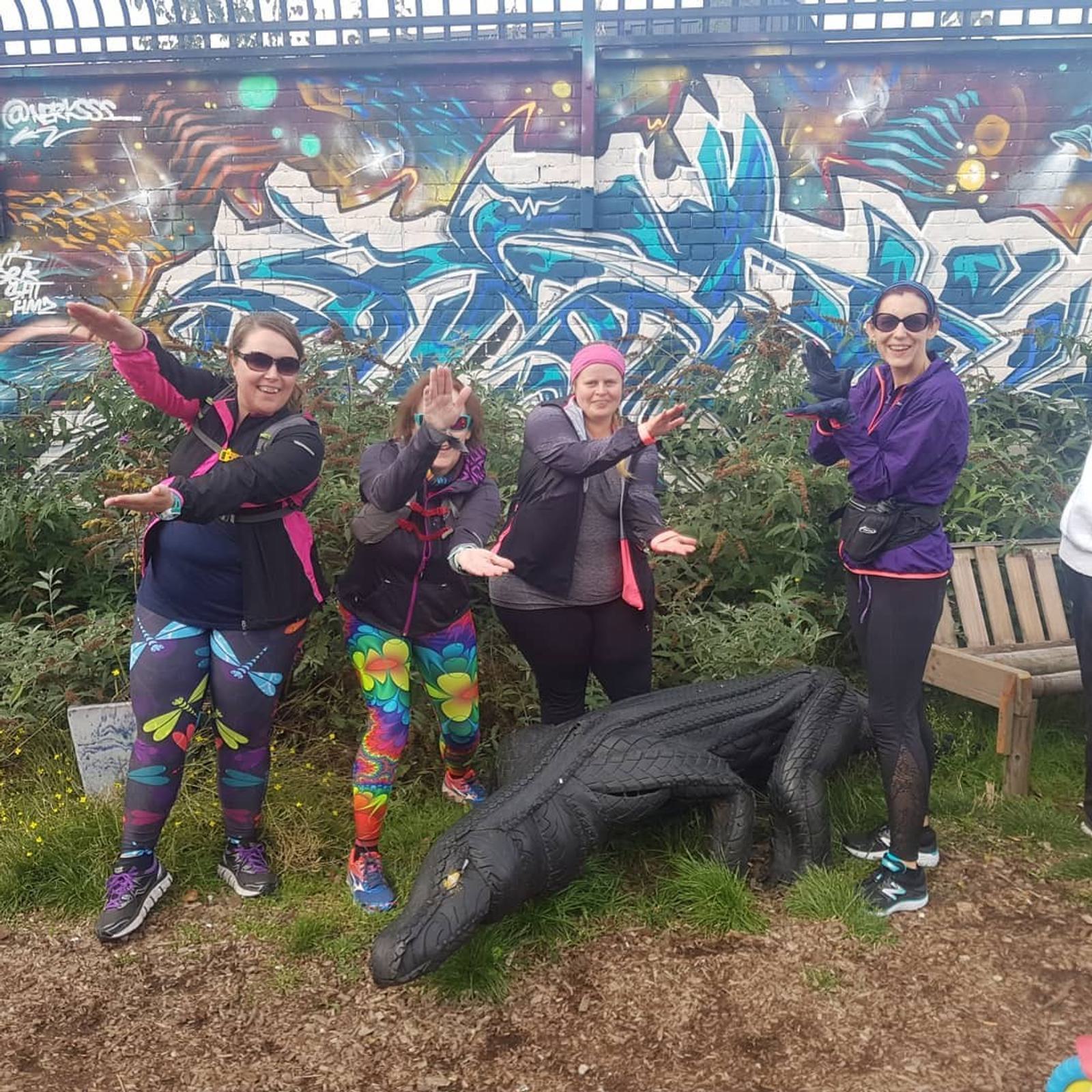 Four smiling adults in colorful athletic wear pose playfully around a black alligator statue. Behind them is a vibrant graffiti mural on a brick wall. Three of them stand, while one kneels, adding a fun and lively atmosphere to the scene.