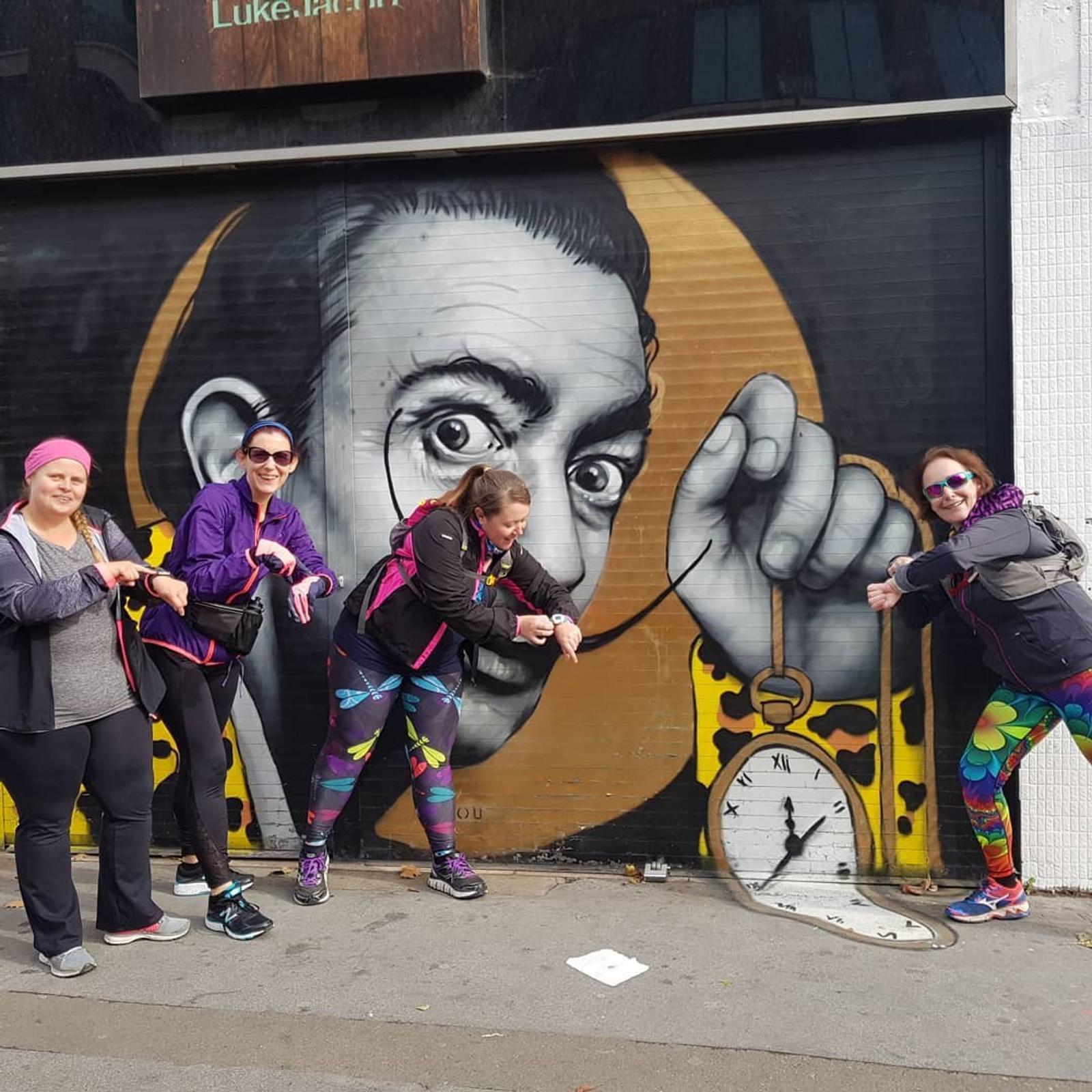 Four women, dressed in colorful athletic wear, pose energetically in front of a street mural featuring a portrait of Salvador Dalí holding a melting clock. The women appear to be playfully interacting with the mural, each striking a unique and lively pose.