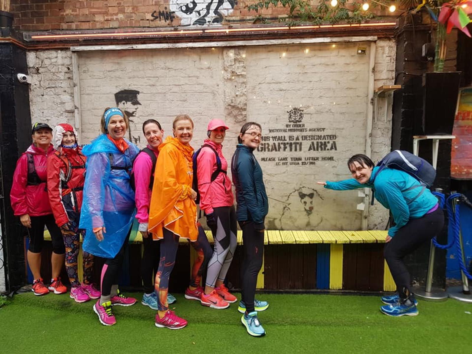 A group of eight people dressed in colorful rain gear pose in front of a mural on a brick wall. The mural announces a designated graffiti area. They seem cheerful, with some pointing at the mural. The area is decorated with graffiti and string lights.