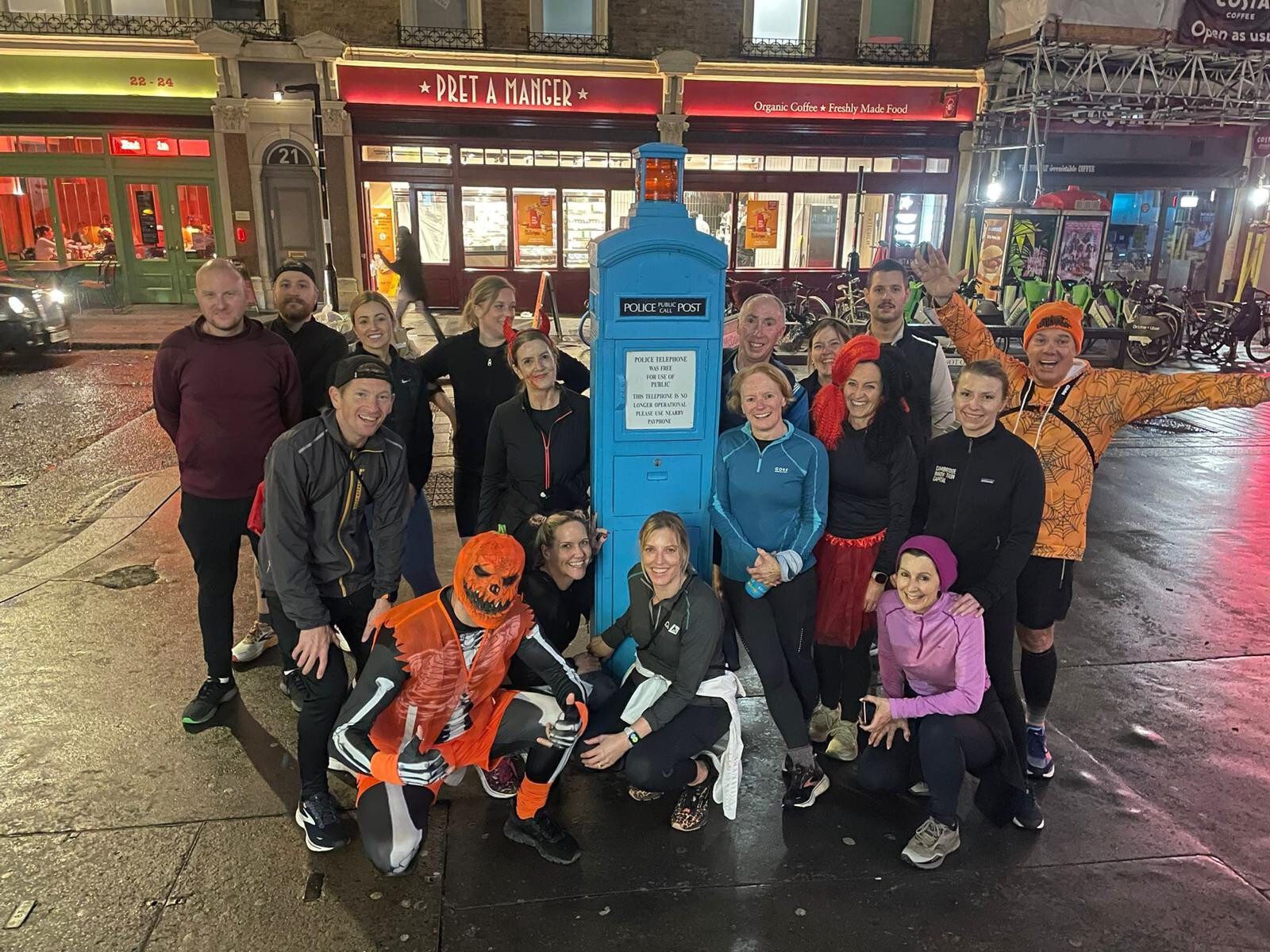 A group of people in colorful outfits, some in costumes, gather around a blue police box on a city street at night. The street is lined with restaurants and a bicycle rack, and the scene is lit by surrounding lights.