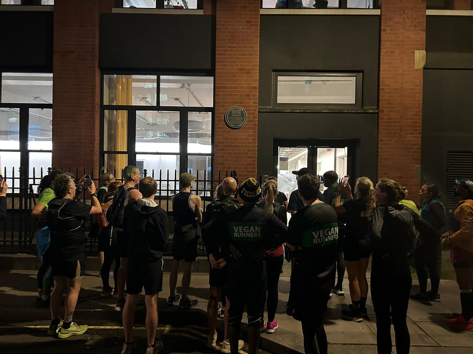 A group of people wearing "Vegan Runners" shirts stand outside a building at night, looking through the windows. The scene is dimly lit, with a brick wall and metal fencing visible in the background.