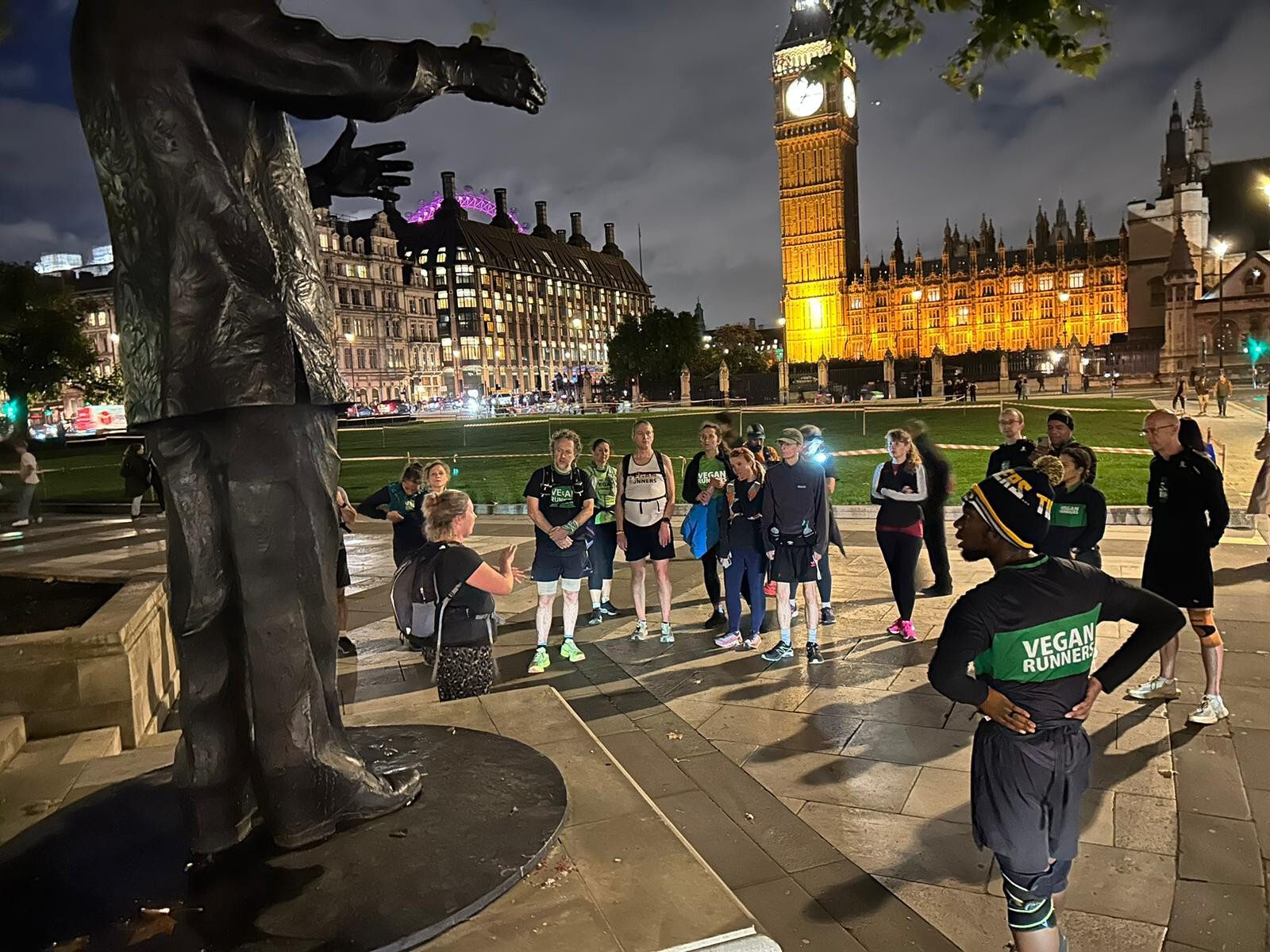A group of people is gathered around a street performer near a statue in an urban area at night. Big Ben and the illuminated Houses of Parliament are in the background, and some people are wearing "Vegan Runner" shirts.