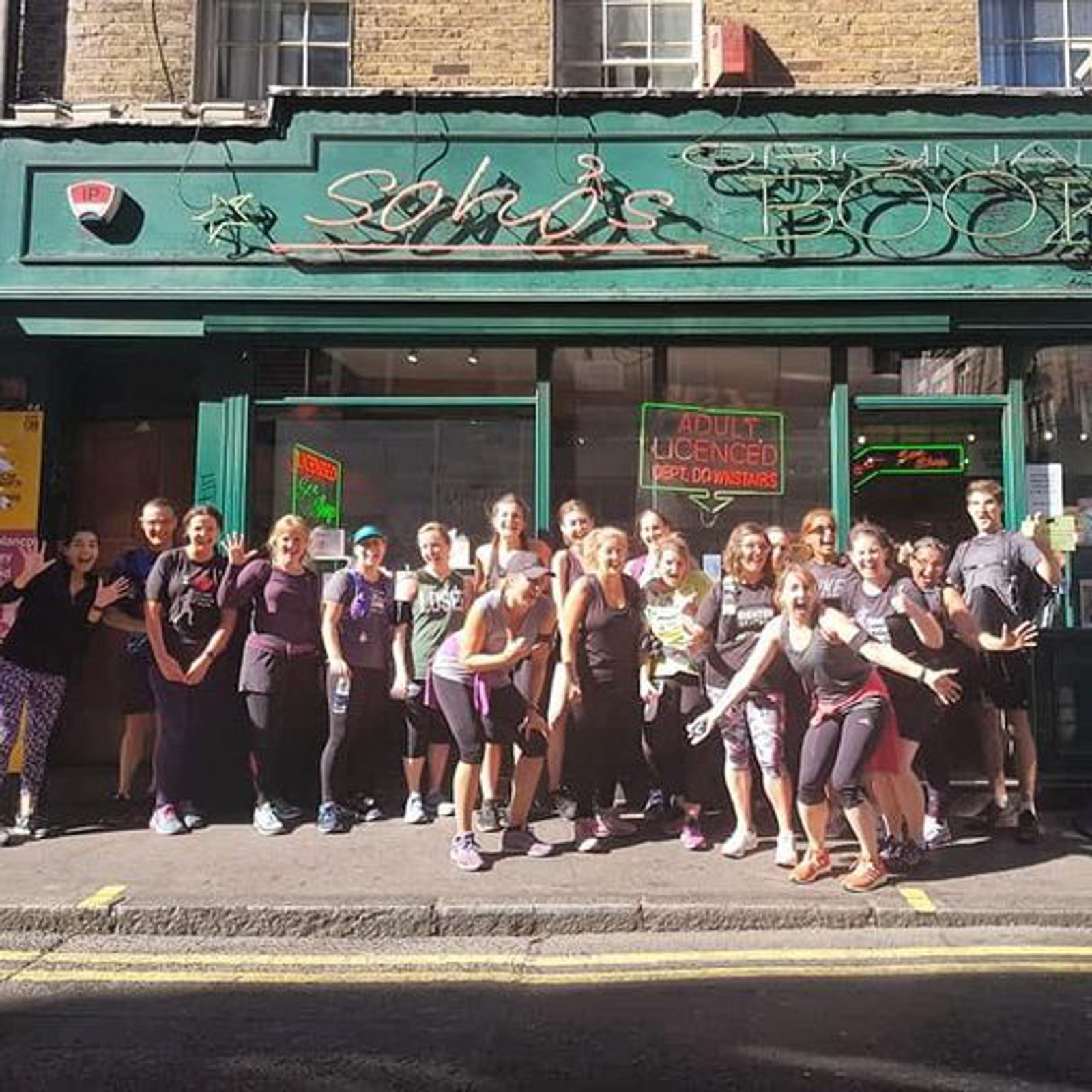 A group of people wearing workout attire are standing and posing in front of a storefront with a green and yellow sign. Some people have their hands raised or are smiling at the camera. The street is sunny, and the storefront has a brick facade.