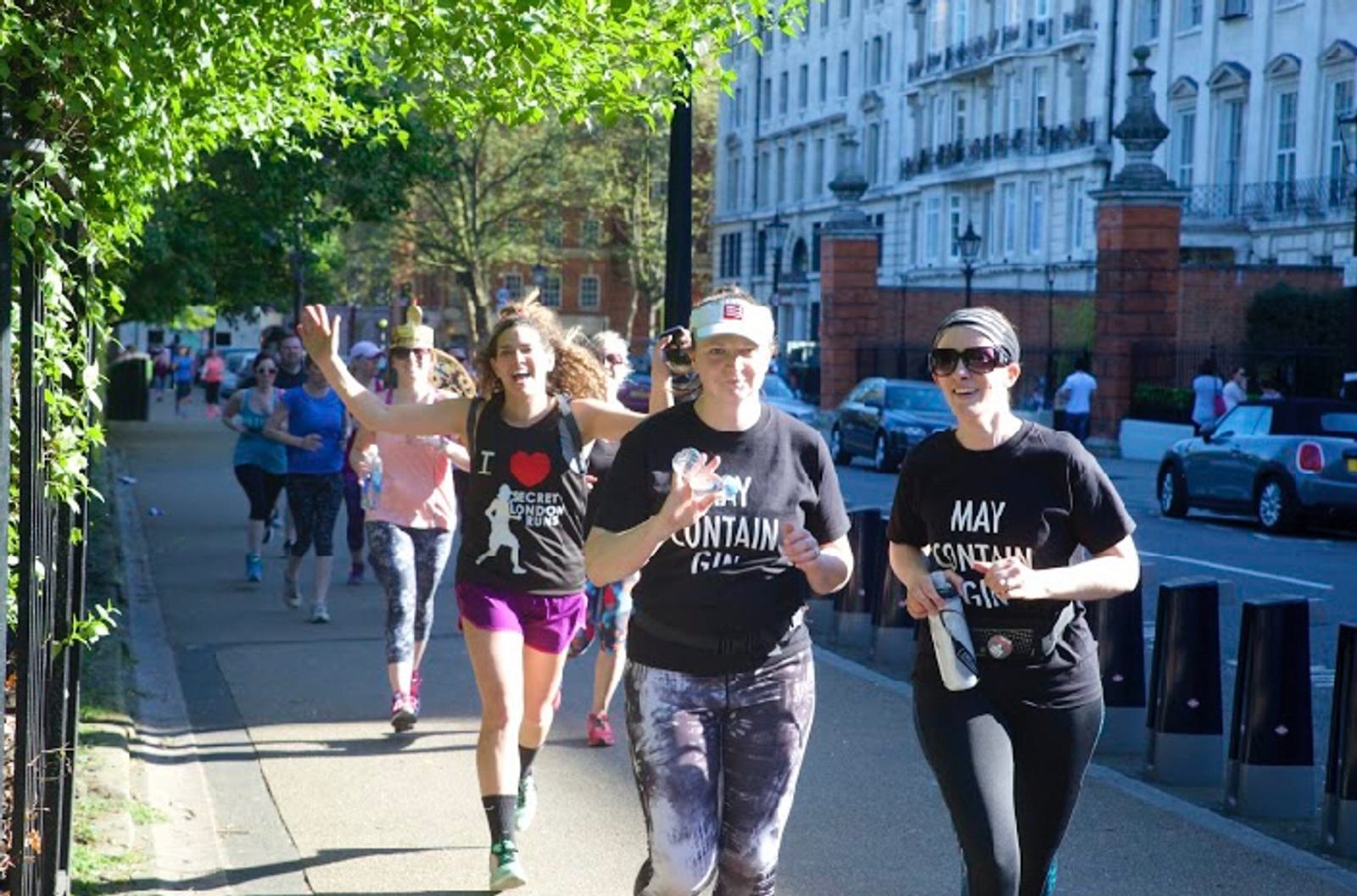 A group of people are running along a tree-lined sidewalk in a city. Two women in the foreground wear black shirts with white text, and one has a paper cup. Another woman behind them waves cheerfully. More runners can be seen in the background.