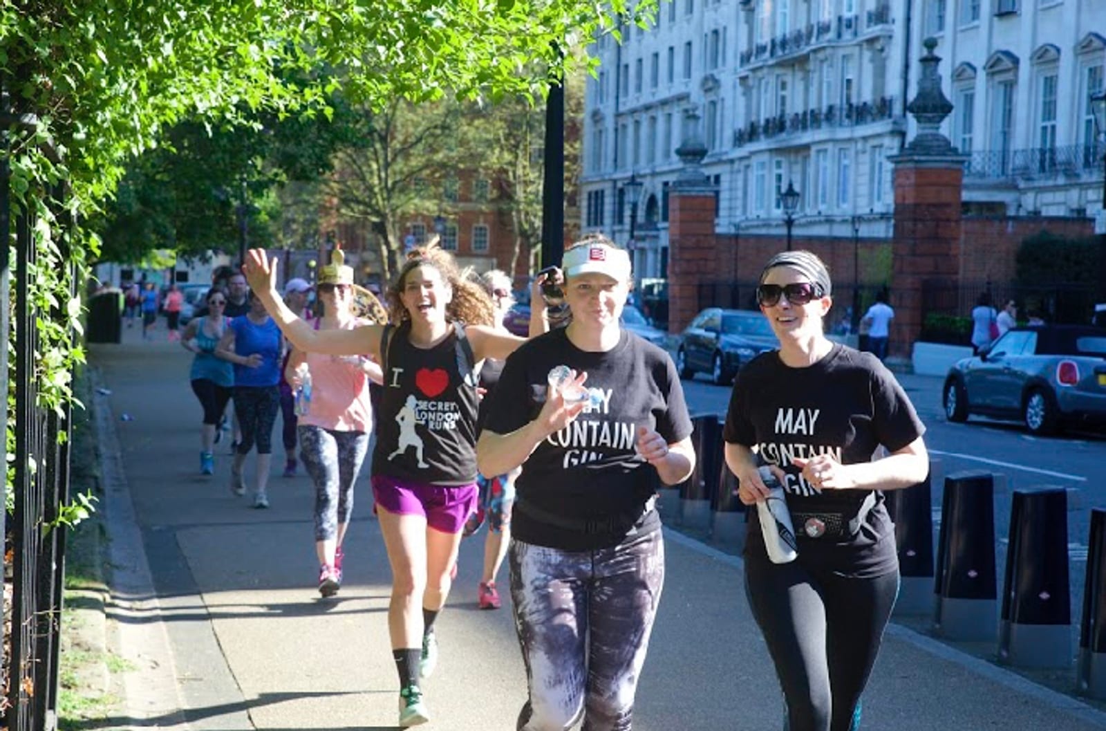 A group of people is participating in a fun run on a sunny day. Three women in the foreground are running and smiling, wearing athletic gear and sunglasses. Other participants are seen in the background along a tree-lined street with buildings.