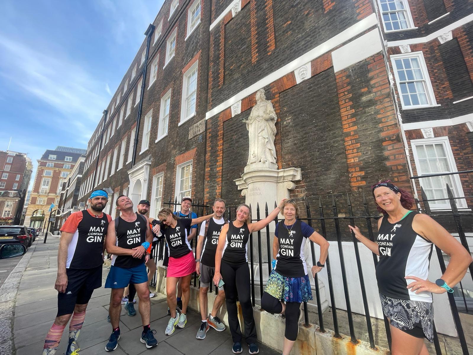 A group of nine people, dressed in running gear and "May Contain Gin" shirts, pose on a city street near a historical building with a statue. They appear happy and energized, standing against a backdrop of red brick architecture and a clear sky.