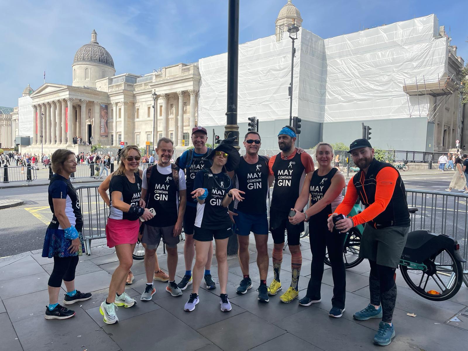 A group of nine runners posing on a street with historic buildings in the background. They are all wearing matching black tank tops with "May Contain Gin" printed on them. Some of them are smiling and making playful gestures. The sky is clear and sunny.