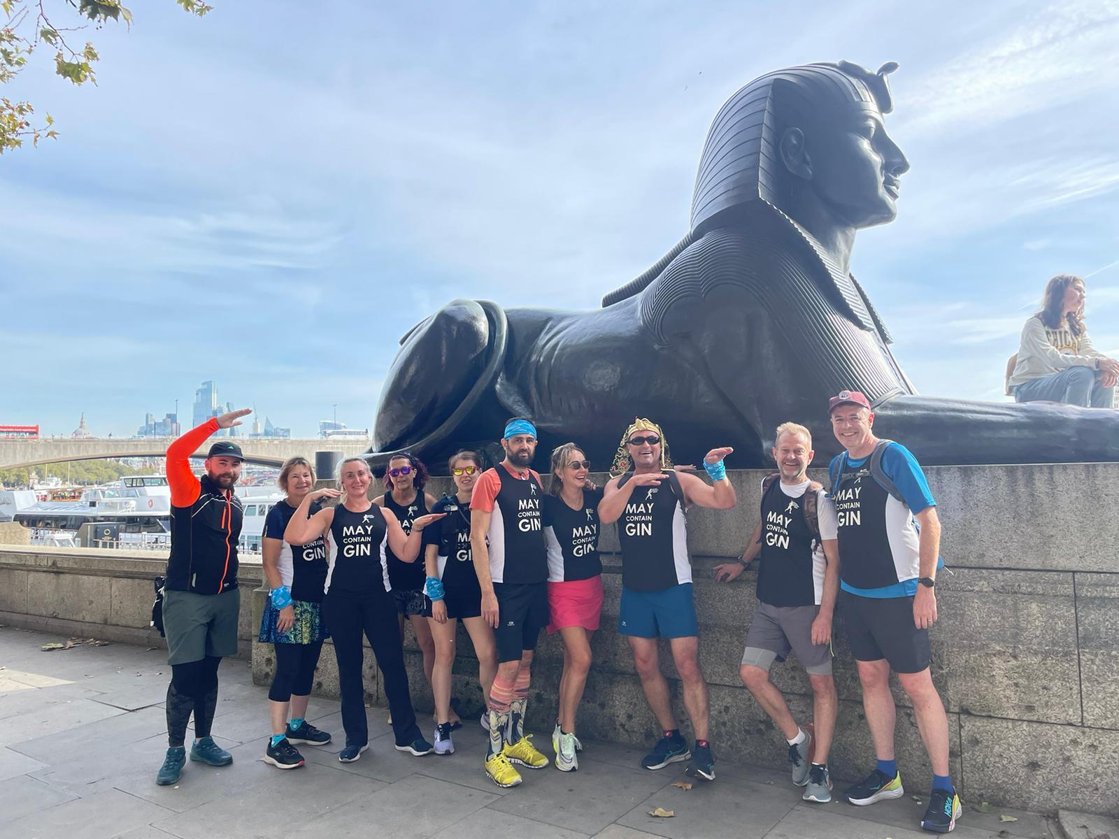 A group of runners pose and smile in front of an Egyptian-style sphinx statue on an embankment. Most are wearing matching tank tops with "MAY CON" written on them. A cityscape and River Thames are visible in the background against a sunny sky.