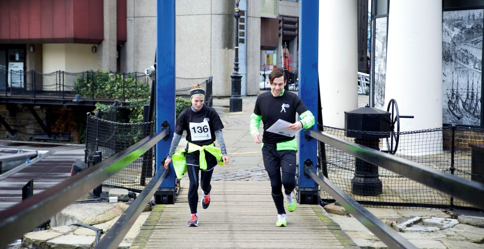 Two people, a man and a woman, dressed in athletic gear, are jogging across a bridge. The woman, wearing a black shirt with the number 136, is slightly ahead, while the man is holding a map. The background features urban buildings and a lamppost.