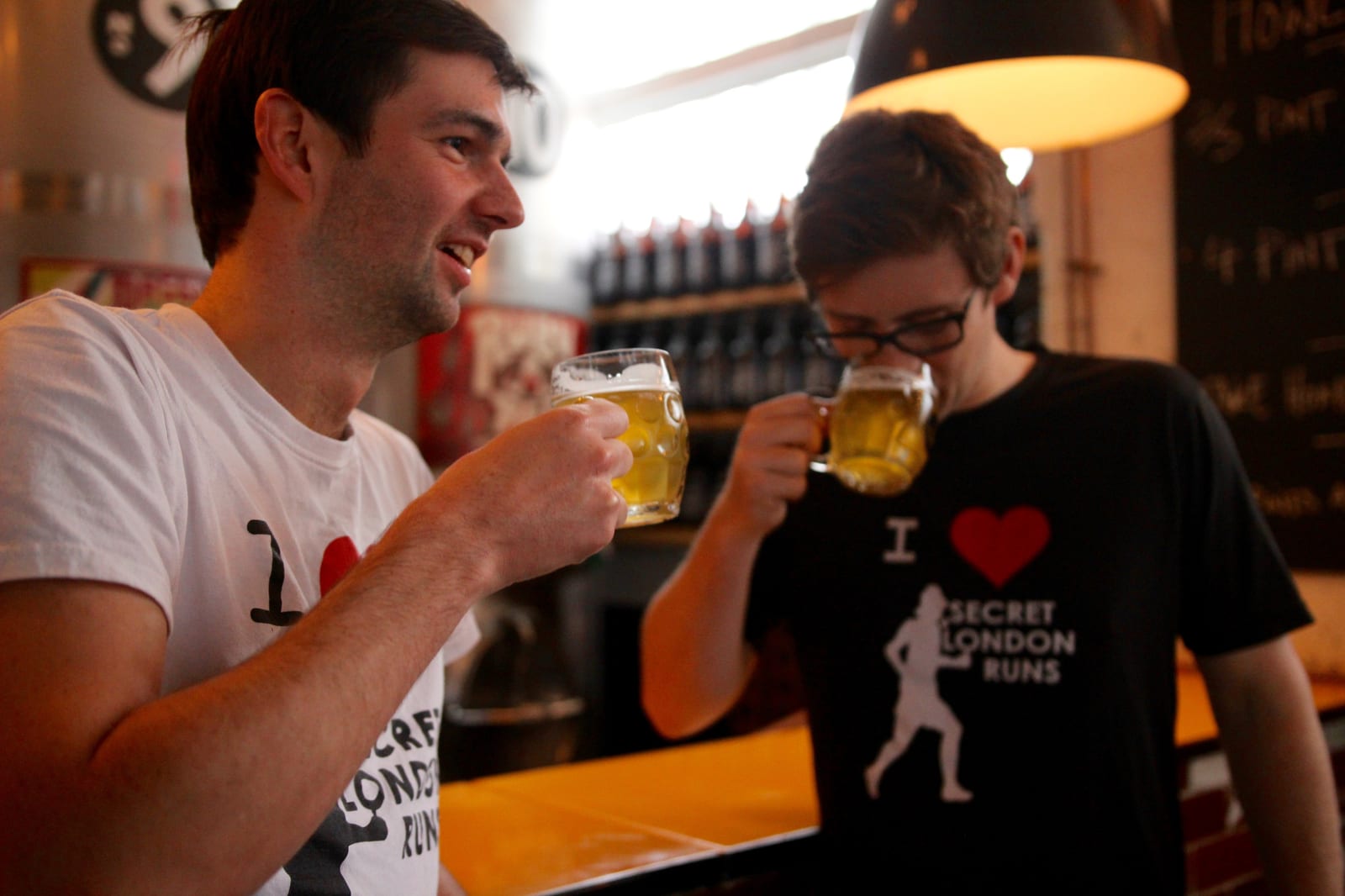 Two men are standing in a bar, drinking beer out of glass mugs. Both are wearing black and white T-shirts with "I love Secret London Runs" on them. One man is taking a sip while the other is smiling and chatting, with a warmly lit bar area behind them.