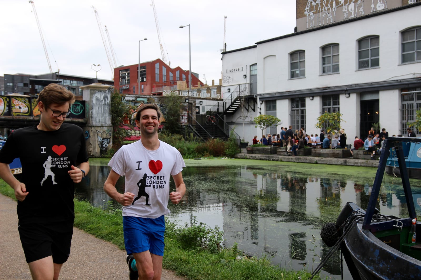 Two people jog along a canal path wearing "I ♥ Secret London Runs" T-shirts. They smile as they run past a mix of modern and older buildings, with a group of people gathered on the opposite canal bank. Cranes and greenery are visible in the background.