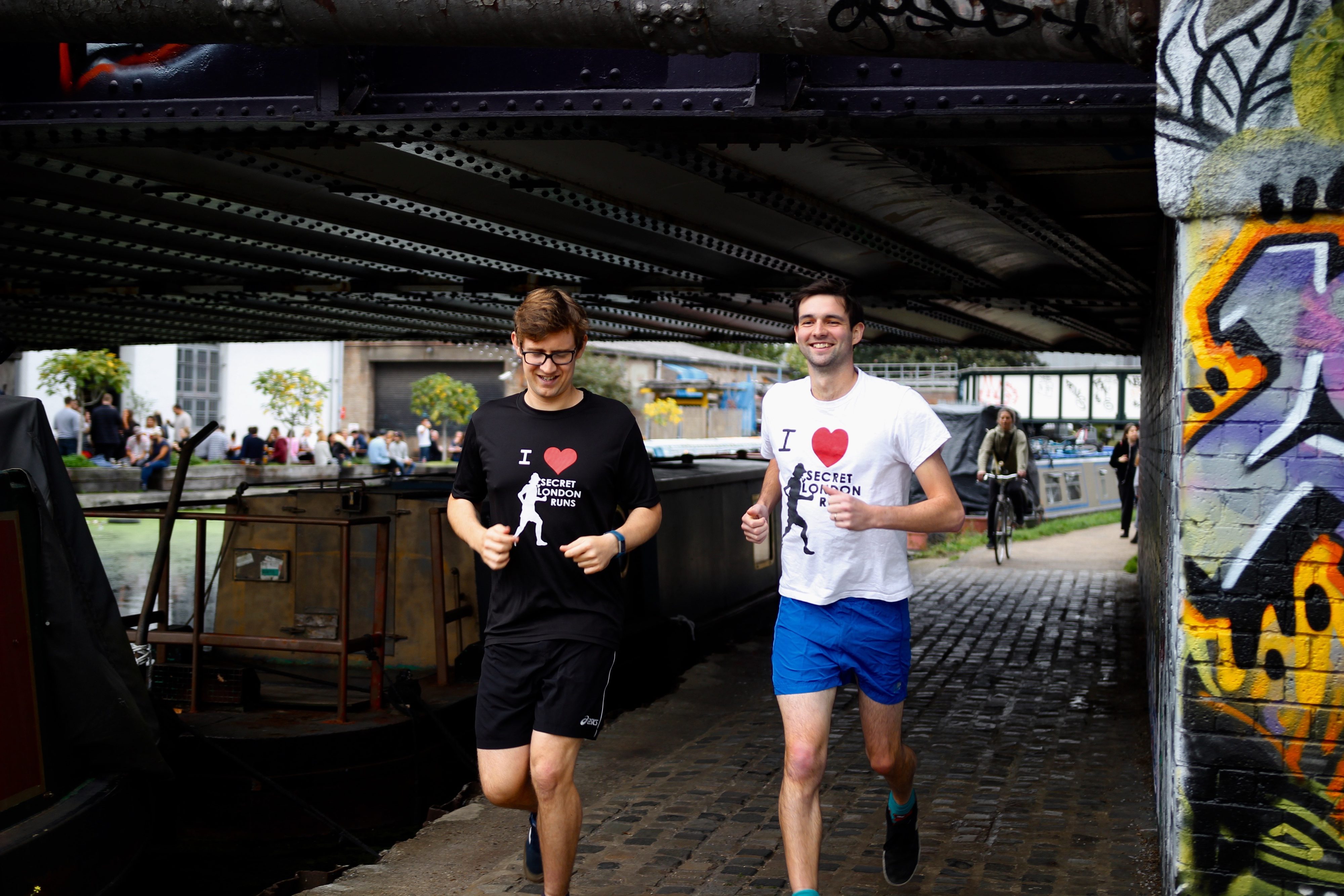 Two men are jogging under a bridge along a waterside path. Both are wearing glasses and t-shirts with "I ♥ London" printed on them. The man on the left is in black shorts and a black shirt, while the man on the right is in blue shorts and a white shirt.