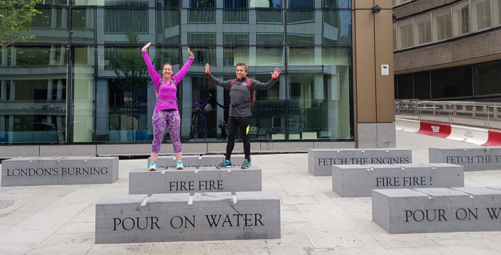 Two people stand on raised concrete blocks with inscriptions like "FIRE FIRE" and "POUR ON WATER" outside a modern glass building. They have their arms raised and are smiling. The sidewalk and part of the street are visible in the background.