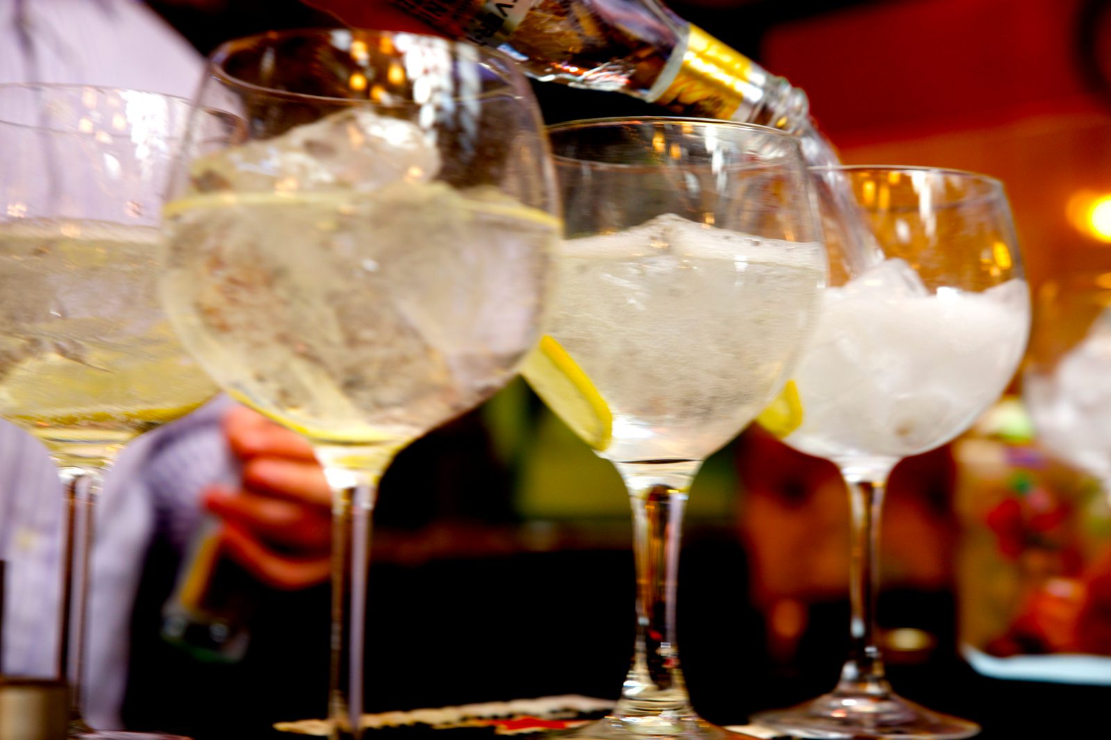 Close-up of several large wine glasses filled with ice and garnished with lemon slices, being filled with a clear liquid from a bottle. The image is brightly lit and appears to be taken in a festive or social setting.