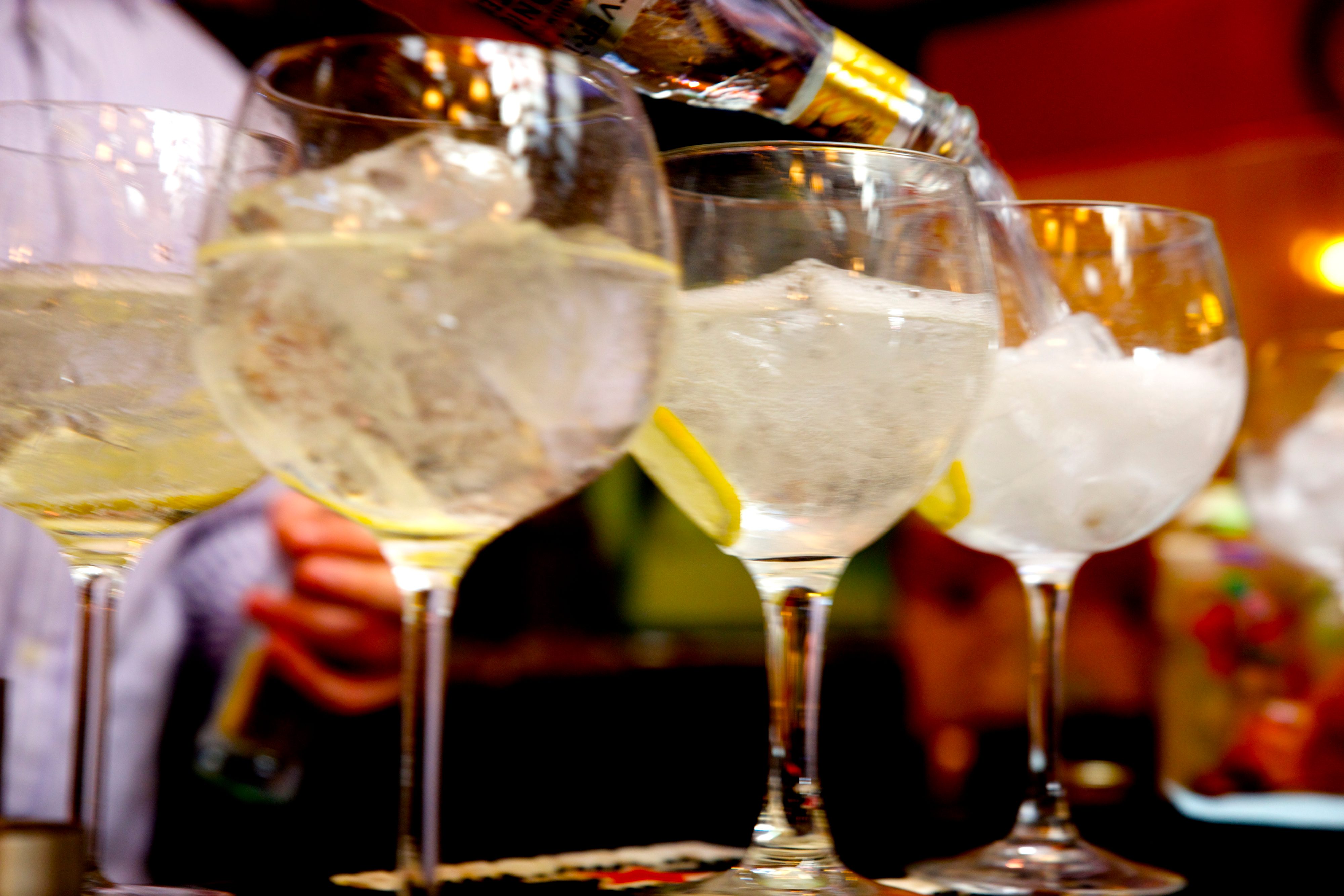 Close-up of several large wine glasses filled with ice and garnished with lemon slices, being filled with a clear liquid from a bottle. The image is brightly lit and appears to be taken in a festive or social setting.