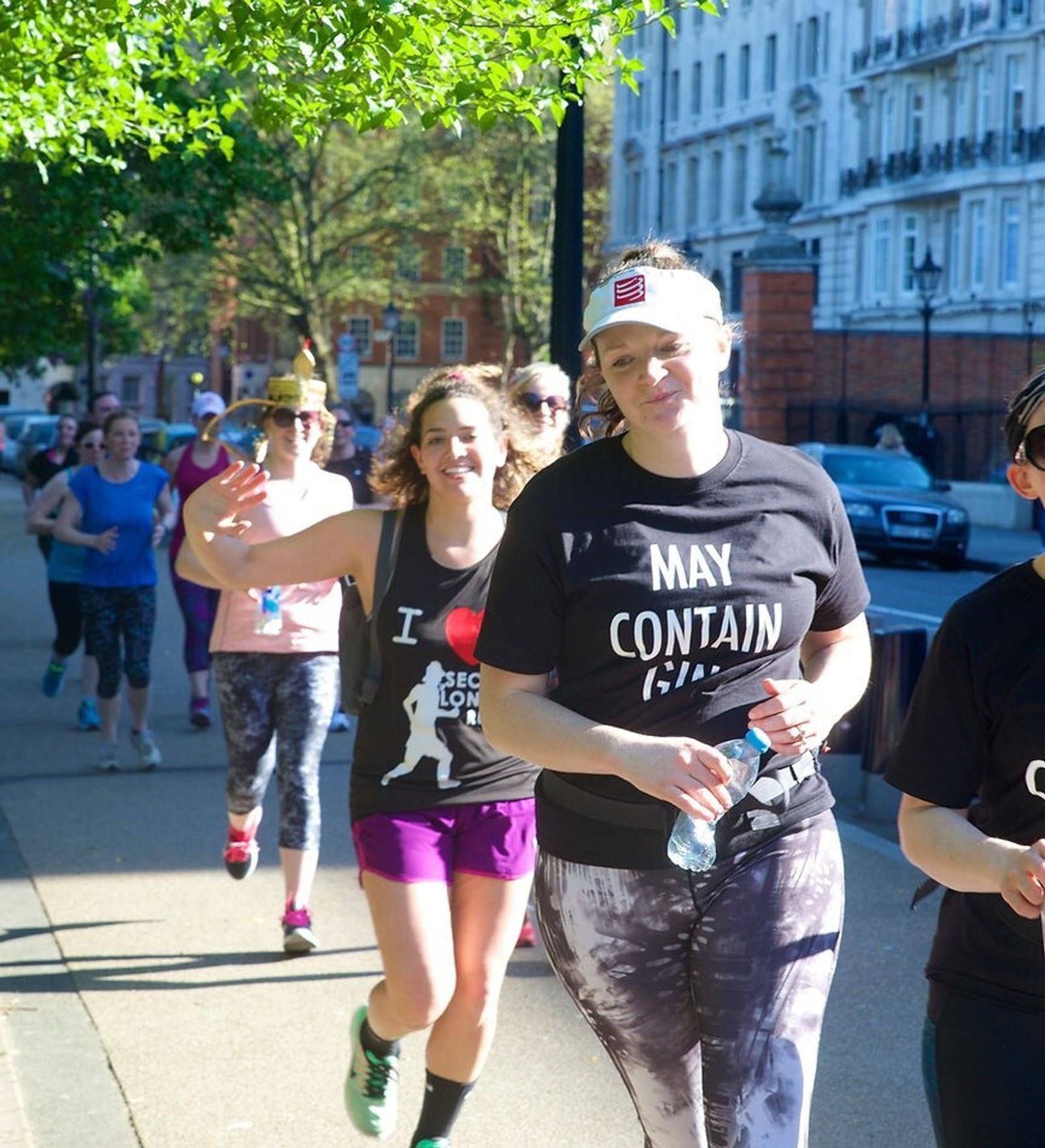 A group of people participating in a lively outdoor run on a sunny day, with trees and buildings in the background. The foreground features a woman in a "May Contain Gin" shirt and another person behind her waving cheerfully at the camera.