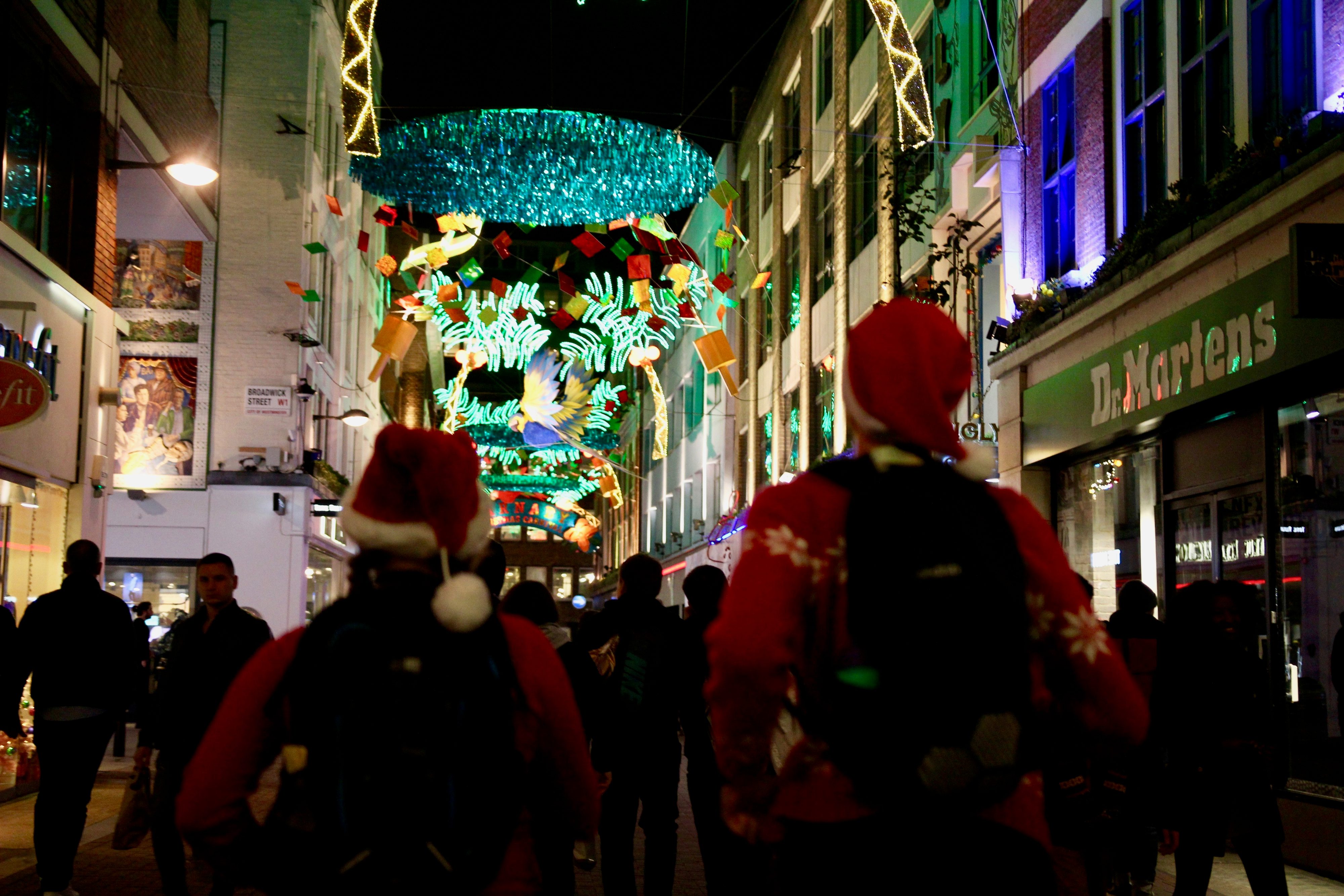 A festive nighttime street scene features people walking under vibrant holiday decorations. Two individuals in the foreground wear Santa hats and red jackets with backpacks. Colorful lights and banners adorn the shop-lined street, contributing to the festive atmosphere.